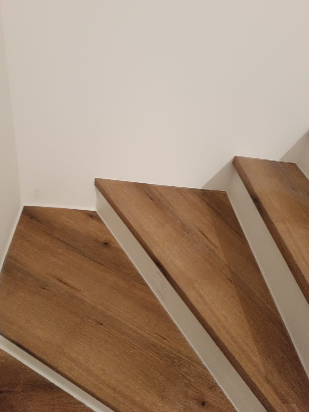 Fragment schodów z drewnopodobnymi panelami i białymi listwami przypodłogowymi, widok z dołu, oświetlenie naturalne.