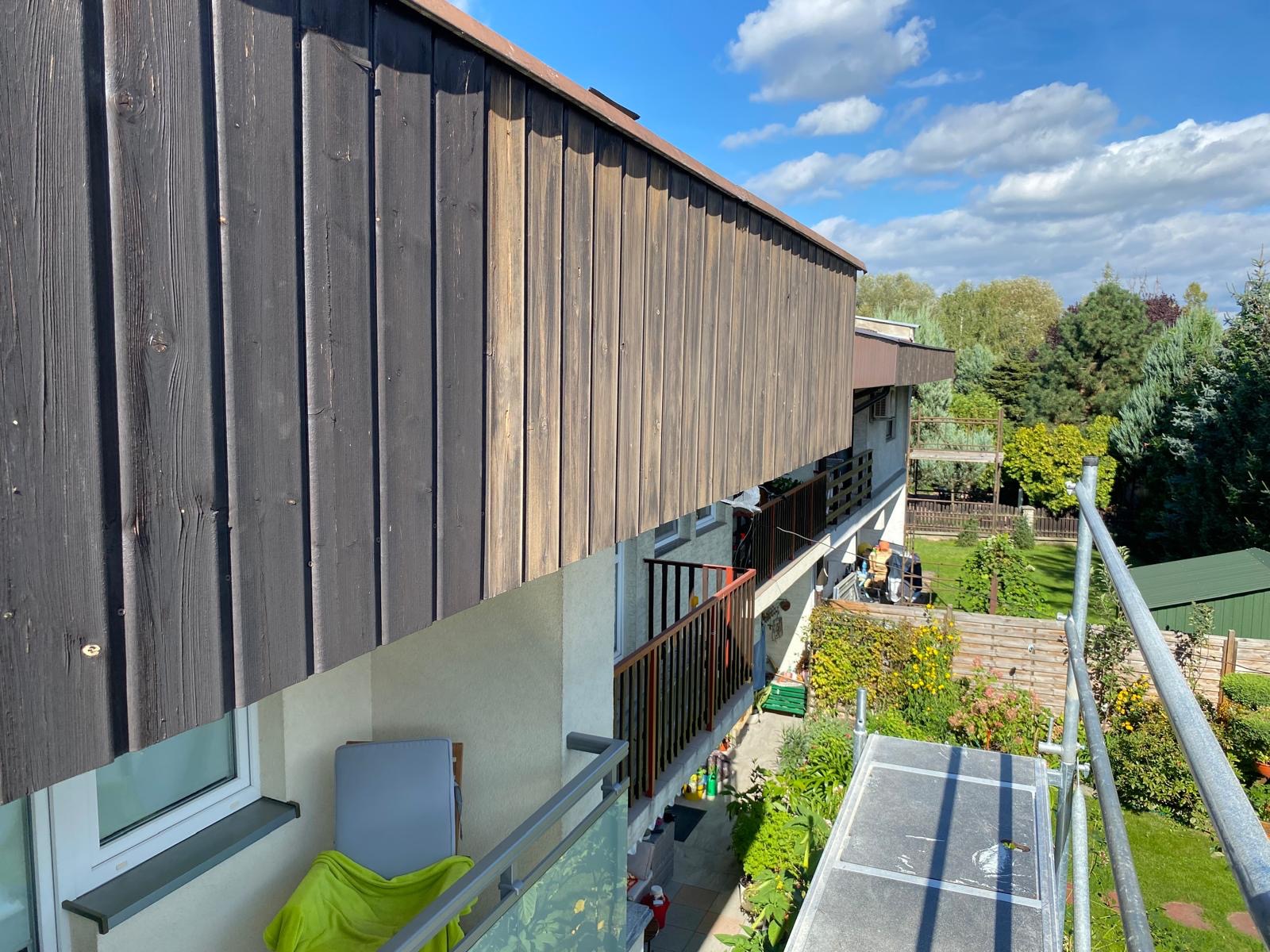 Odnowiona drewniana elewacja balkonu z ciemnymi i jasnymi deskami, widok z rusztowania na tle zieleni i błękitnego nieba z chmurami.