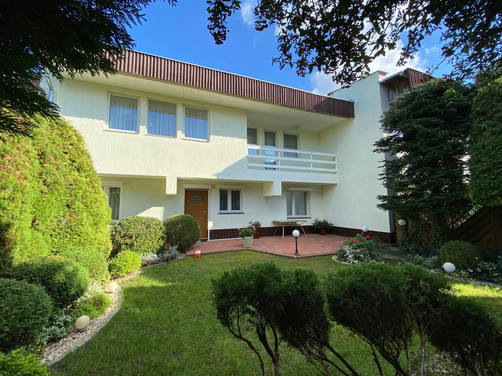 Dwupiętrowy dom z balkonem w Kraszewicach, kremowa elewacja, brązowe akcenty, zadbany ogród z krzewami i trawnikiem. Widok z poziomu gruntu, słoneczny dzień.