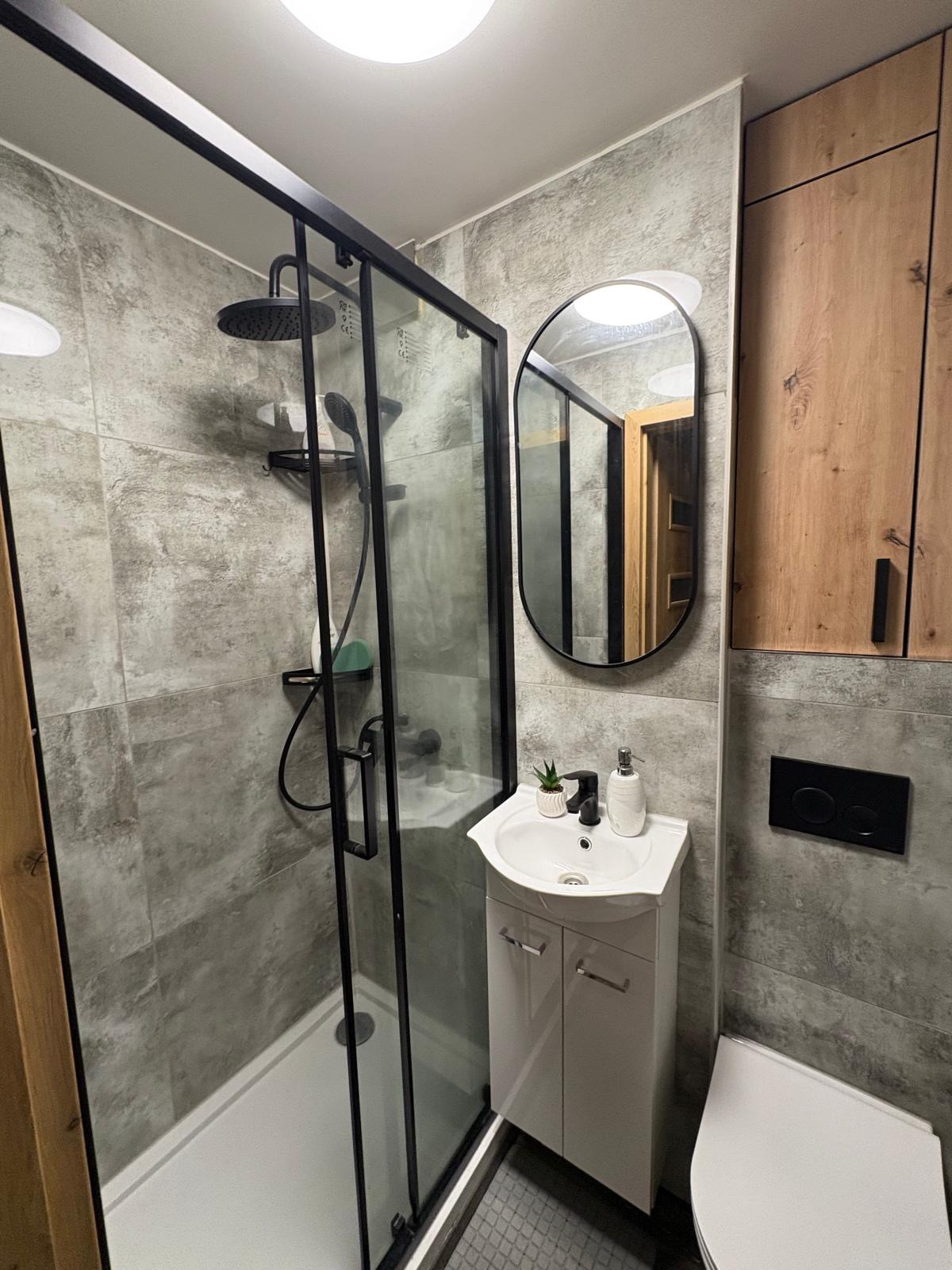 Nowoczesna łazienka z kabiną prysznicową typu walk-in, umywalką z szafką i lustrem w czarnej ramie. Szare płytki imitujące beton i drewniane szafki dodają elegancji.