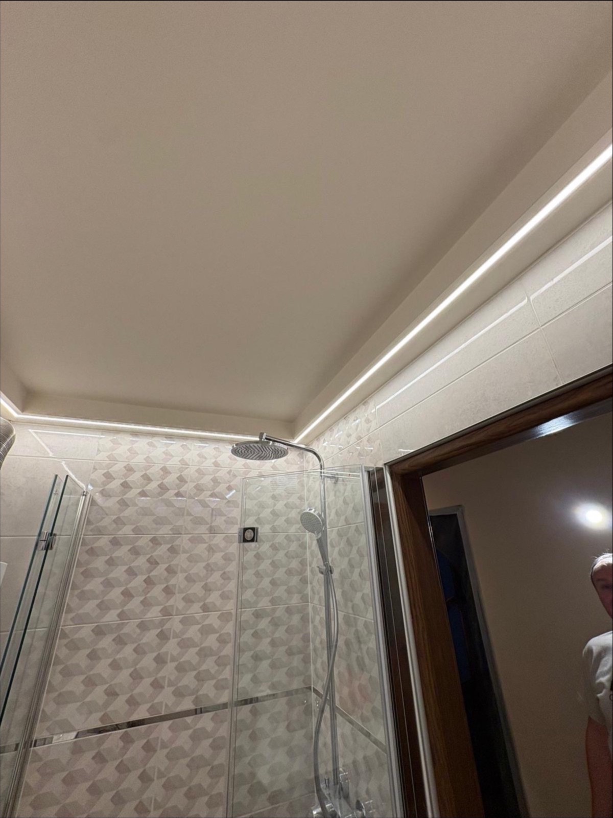 Nowoczesna kabina prysznicowa z geometrycznymi płytkami, oświetleniem LED podsufitowym i szklanymi drzwiami, widziana pod kątem z dołu.