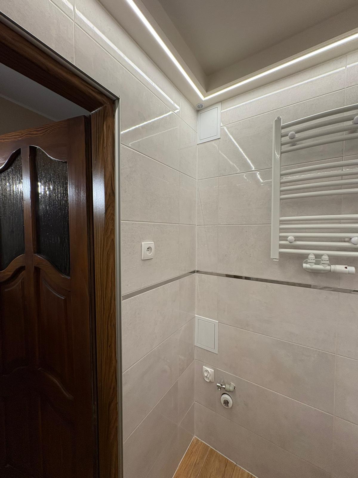 Beżowe płytki w łazience z drewnianą podłogą i grzejnikiem drabinkowym. Oświetlenie LED wzdłuż sufitu. Widoczny fragment ciemnych drzwi z szybą.