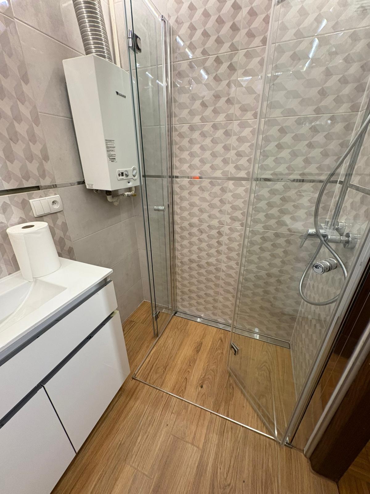 Nowoczesna łazienka po remoncie: kabina prysznicowa ze szklanymi drzwiami, drewnopodobna podłoga, biała szafka pod umywalką i geometryczne płytki na ścianach.