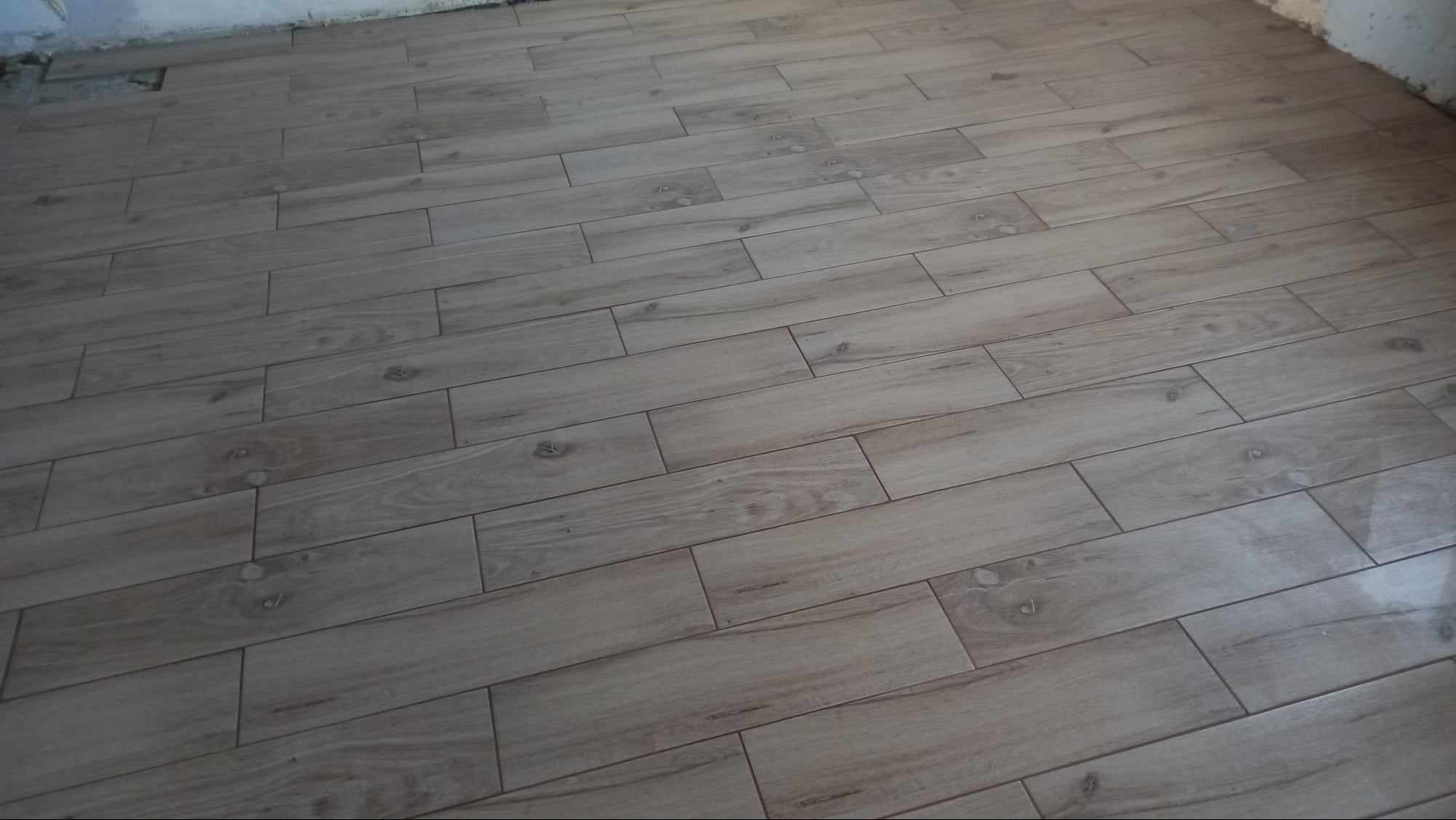 Ułożone, podłużne płytki podłogowe imitujące drewno, z widocznymi fugami. Płytki w odcieniach beżu i brązu, ułożone równolegle do ściany. Widoczny fragment ściany.