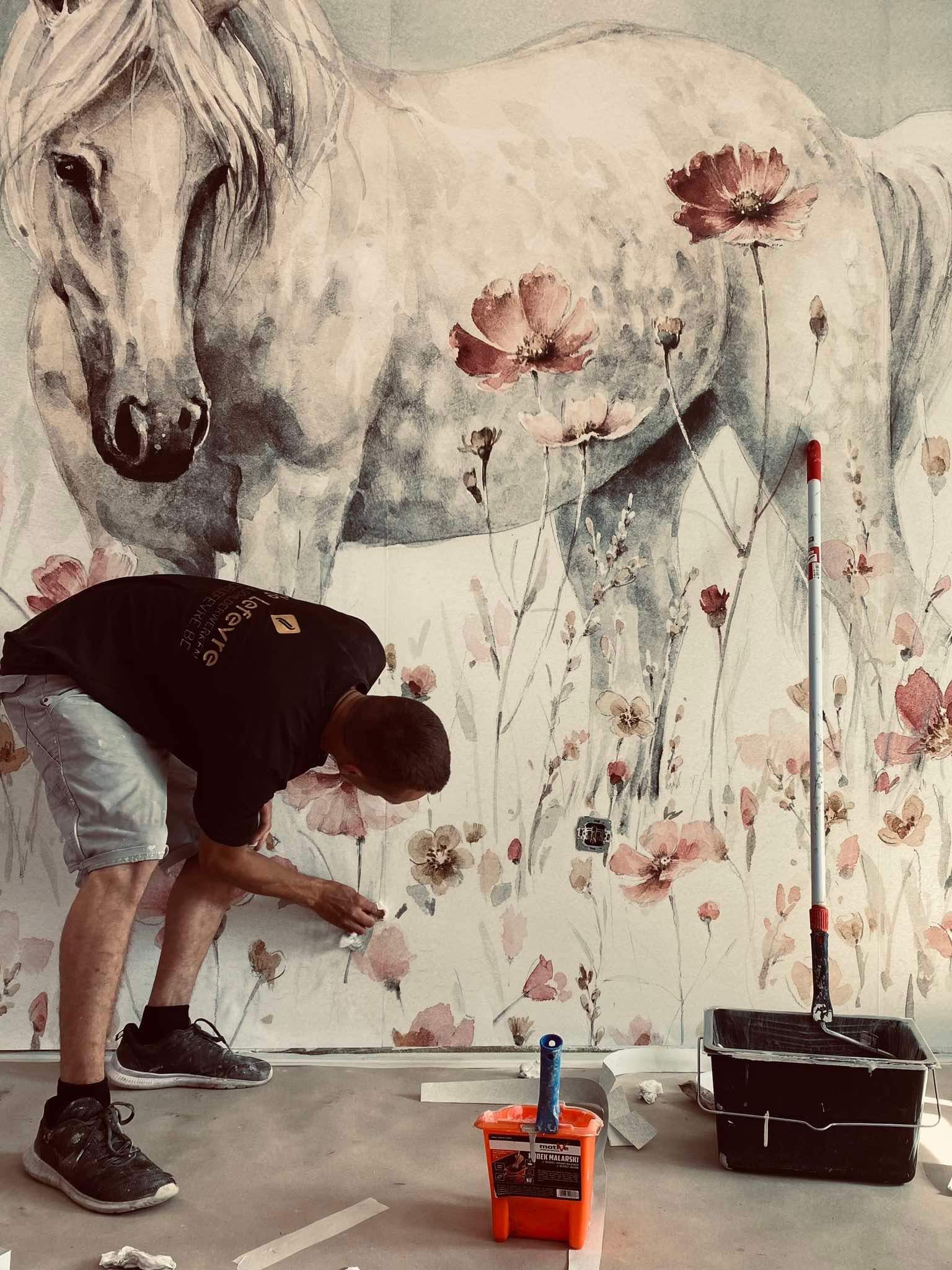 Pracownik wygładza tapetę z motywem konia i kwiatów na ścianie. Widoczne narzędzia malarskie: wałek, kuweta, pędzel. Detal: gniazdko elektryczne wkomponowane w wzór.