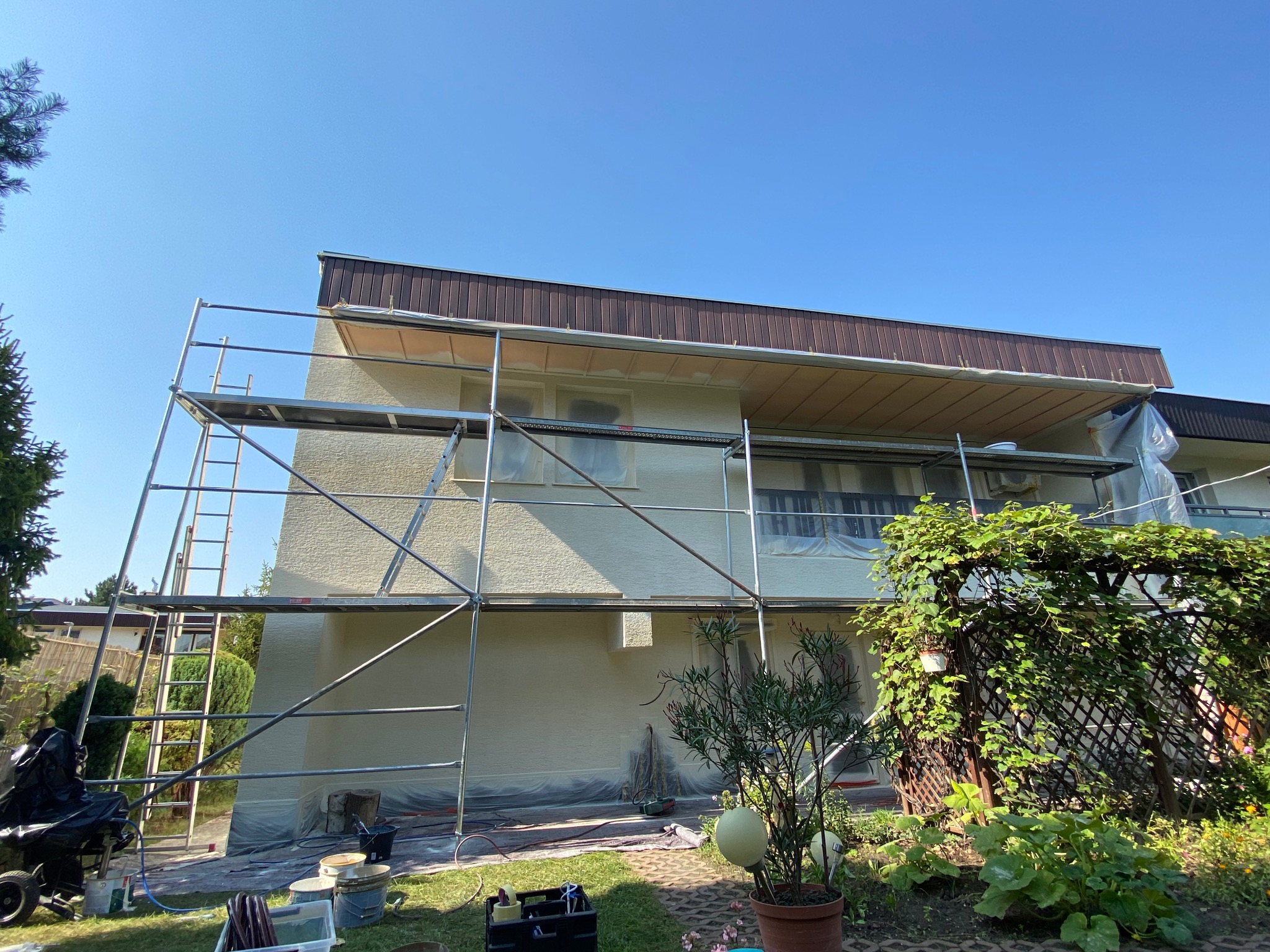 Elewacja domu w trakcie malowania, rusztowanie, zabezpieczone okna folią, widoczne narzędzia malarskie, słoneczny dzień.