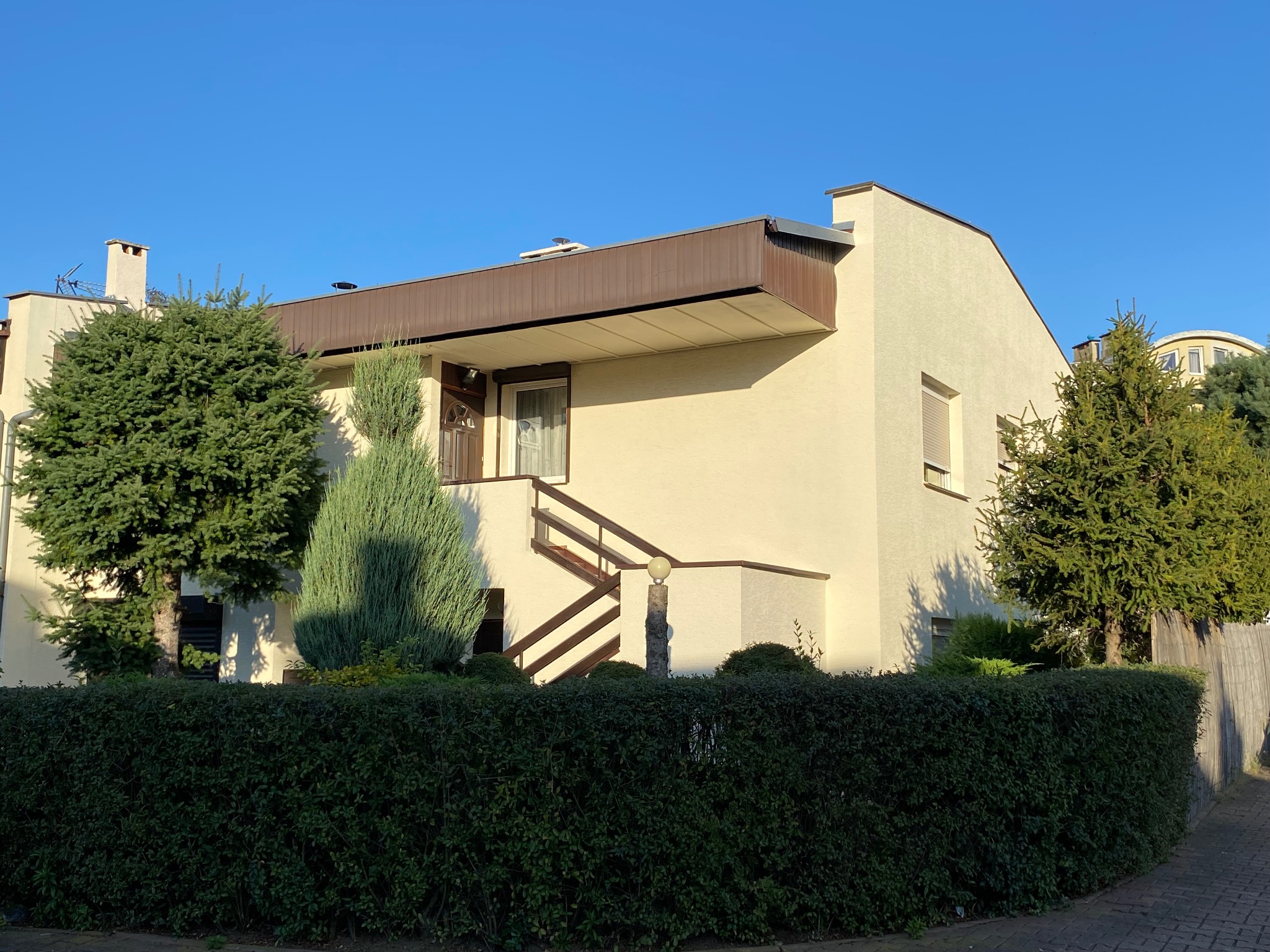 Jasny, dwukondygnacyjny dom z brązowym zadaszeniem nad balkonem i drewnianymi schodami wejściowymi, otoczony zielenią w słoneczny dzień. Widok z poziomu ulicy.