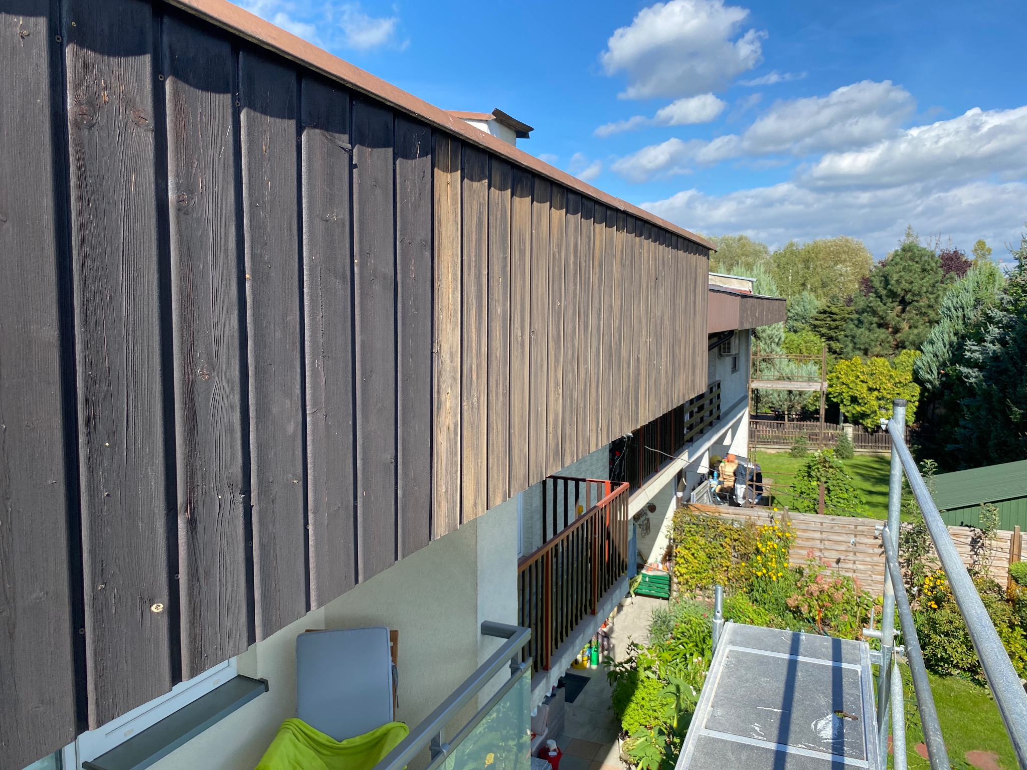 Elewacja budynku z drewnianą okładziną, widok z balkonu. Na drugim planie rusztowanie i zieleń ogrodu. Błękitne niebo z chmurami w tle.