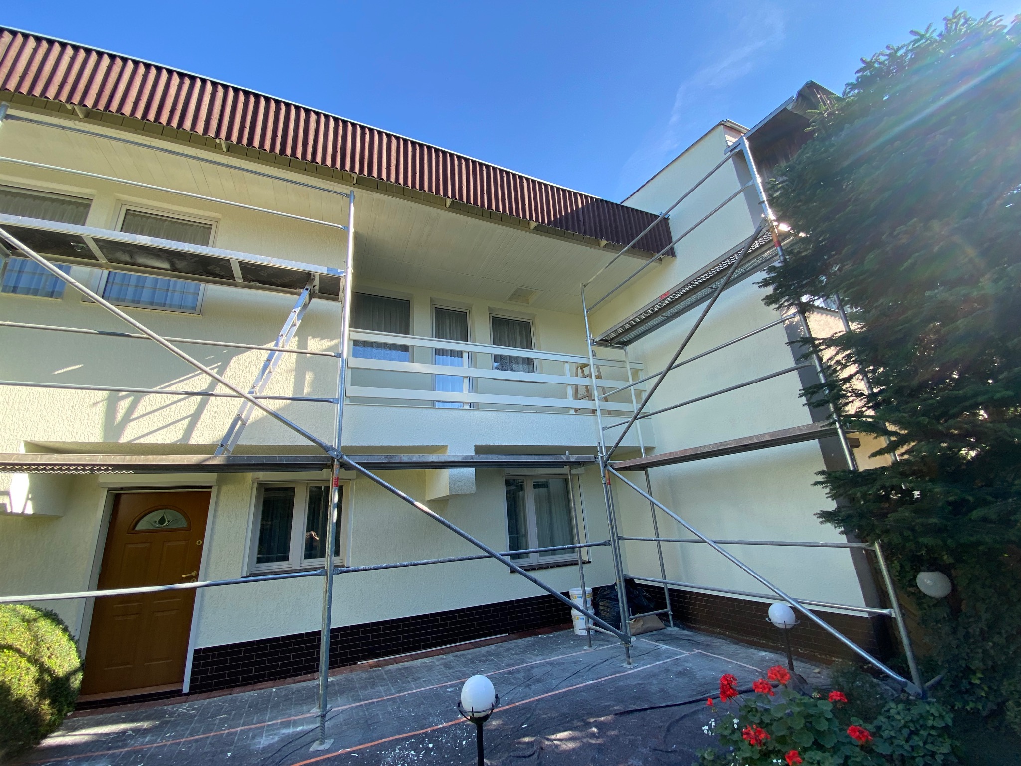 Dom jednorodzinny w trakcie odświeżania elewacji, otoczony rusztowaniem. Widoczne świeżo pomalowane ściany w jasnym odcieniu i elementy zabezpieczające teren prac.