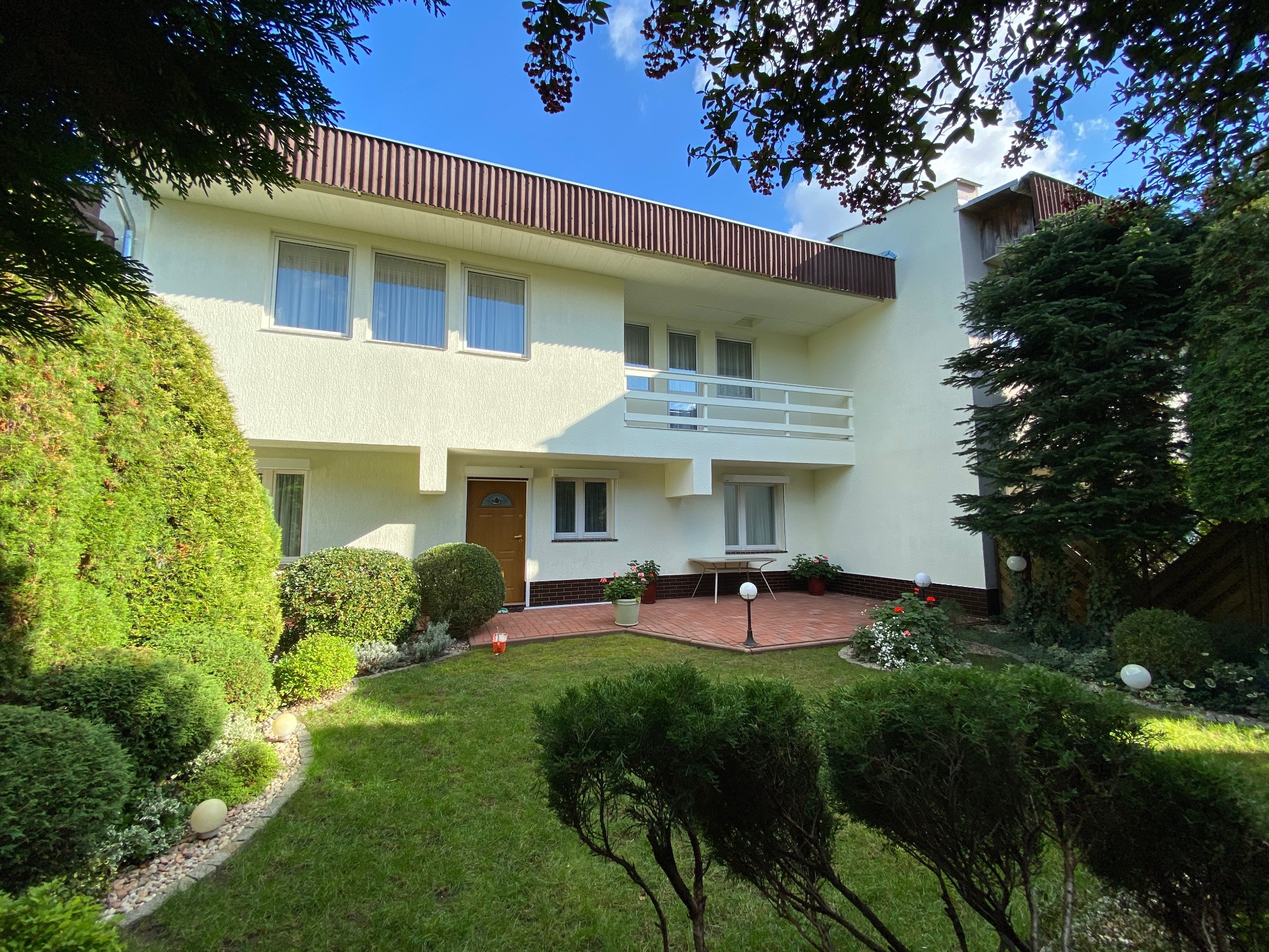 Dwupiętrowy dom z jasną elewacją, balkonem i brązowym dachem, otoczony zielonym ogrodem z krzewami i trawnikiem. Widok z poziomu trawnika, słoneczny dzień.