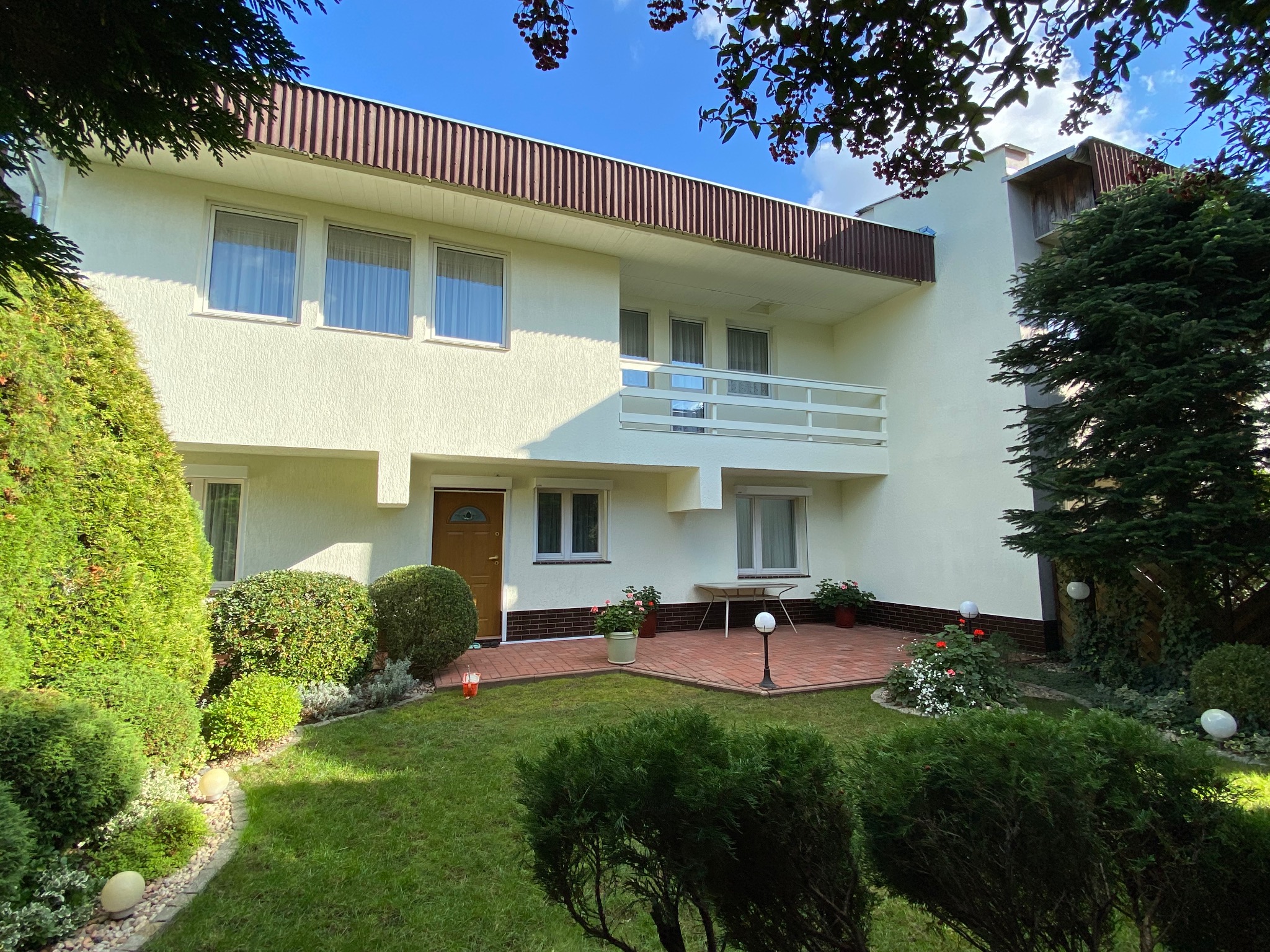 Dwupiętrowy dom z balkonem i brązowym dachem, otoczony zielonym ogrodem z krzewami i trawnikiem. Jasna elewacja i drewniane drzwi wejściowe.