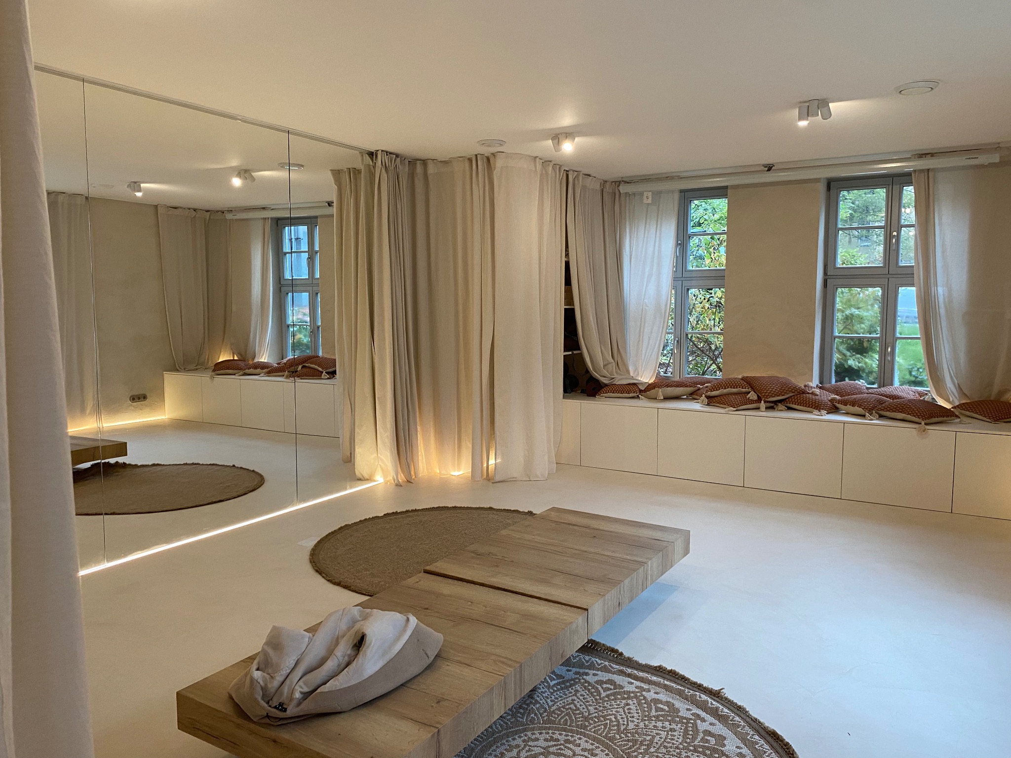 Jasne, minimalistyczne wnętrze w Kraszewicach z drewnianym stolikiem, okrągłymi dywanami i zasłonami. Ściany w odcieniach beżu, duże okna z widokiem na zieleń.