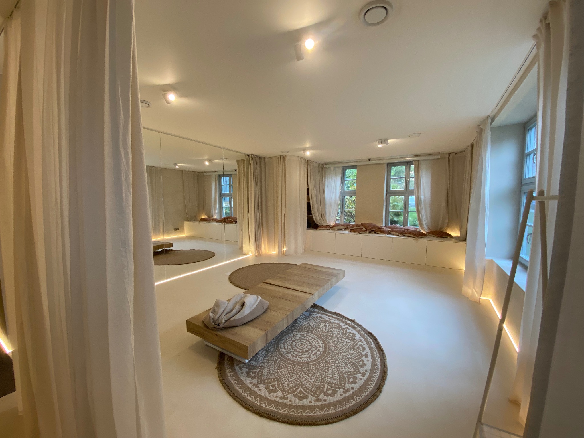 Jasne, przestronne wnętrze z drewnianym stolikiem na okrągłym dywanie, białymi zasłonami i oknami z widokiem na zieleń. Oświetlenie LED podkreśla minimalistyczny design.