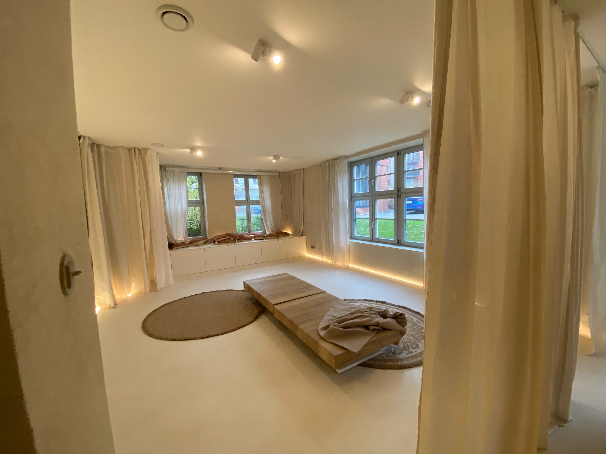 Jasne, minimalistyczne wnętrze z drewnianym podestem, okrągłymi dywanami i zwiewnymi zasłonami. Subtelne oświetlenie LED podkreśla nowoczesny charakter pomieszczenia.