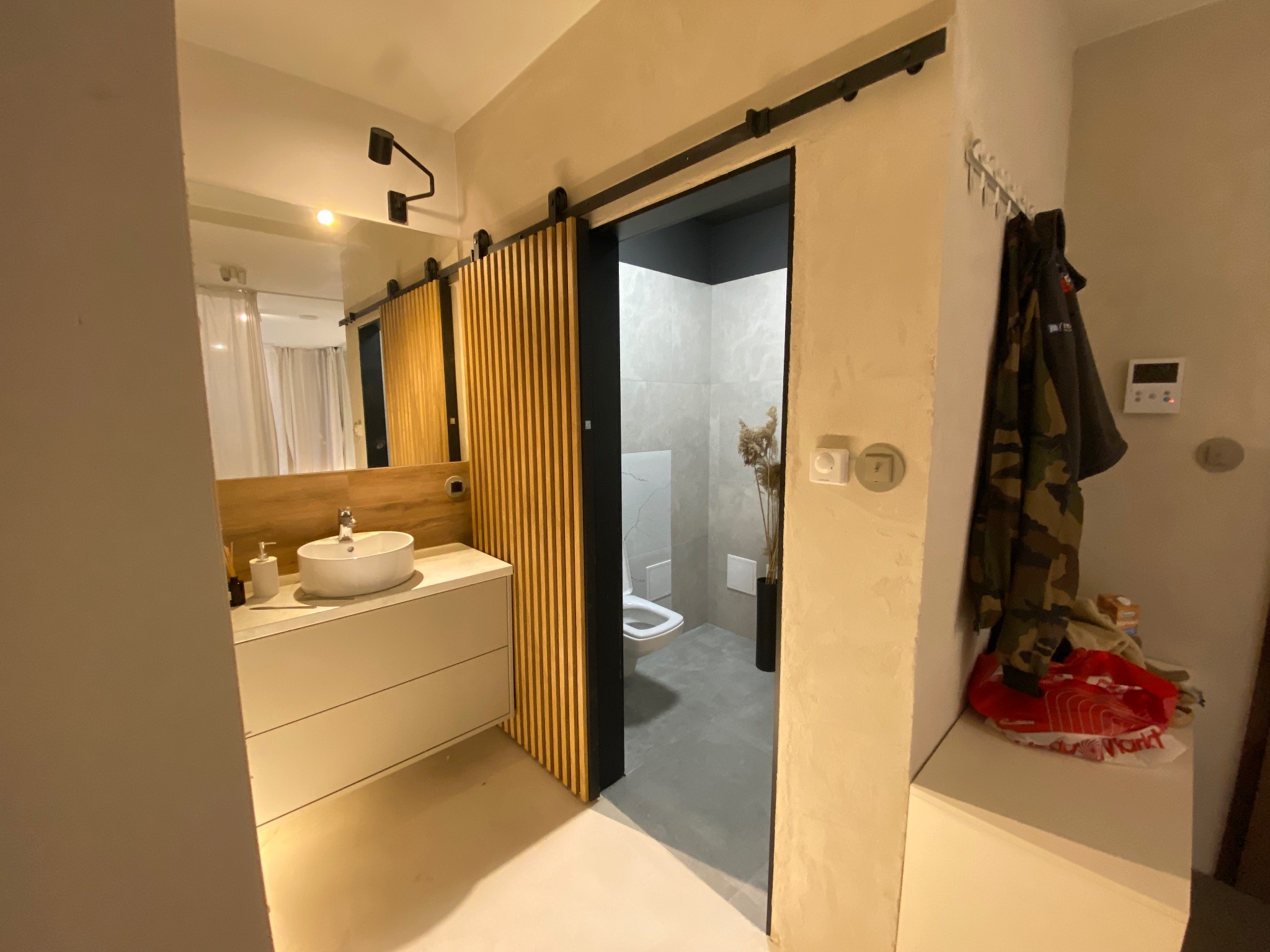 Nowoczesna łazienka z przesuwnymi drzwiami z drewnianych listew, umywalką na szafce i widokiem na toaletę w szaro-białej kolorystyce. Minimalistyczny design.