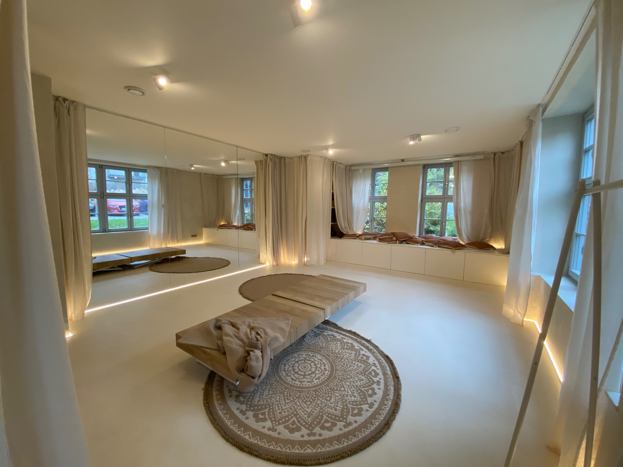 Jasne, przestronne wnętrze z lustrami, drewnianą ławką i okrągłymi dywanami. Subtelne oświetlenie LED podkreśla minimalistyczny design. Widok z okna na zieleń.