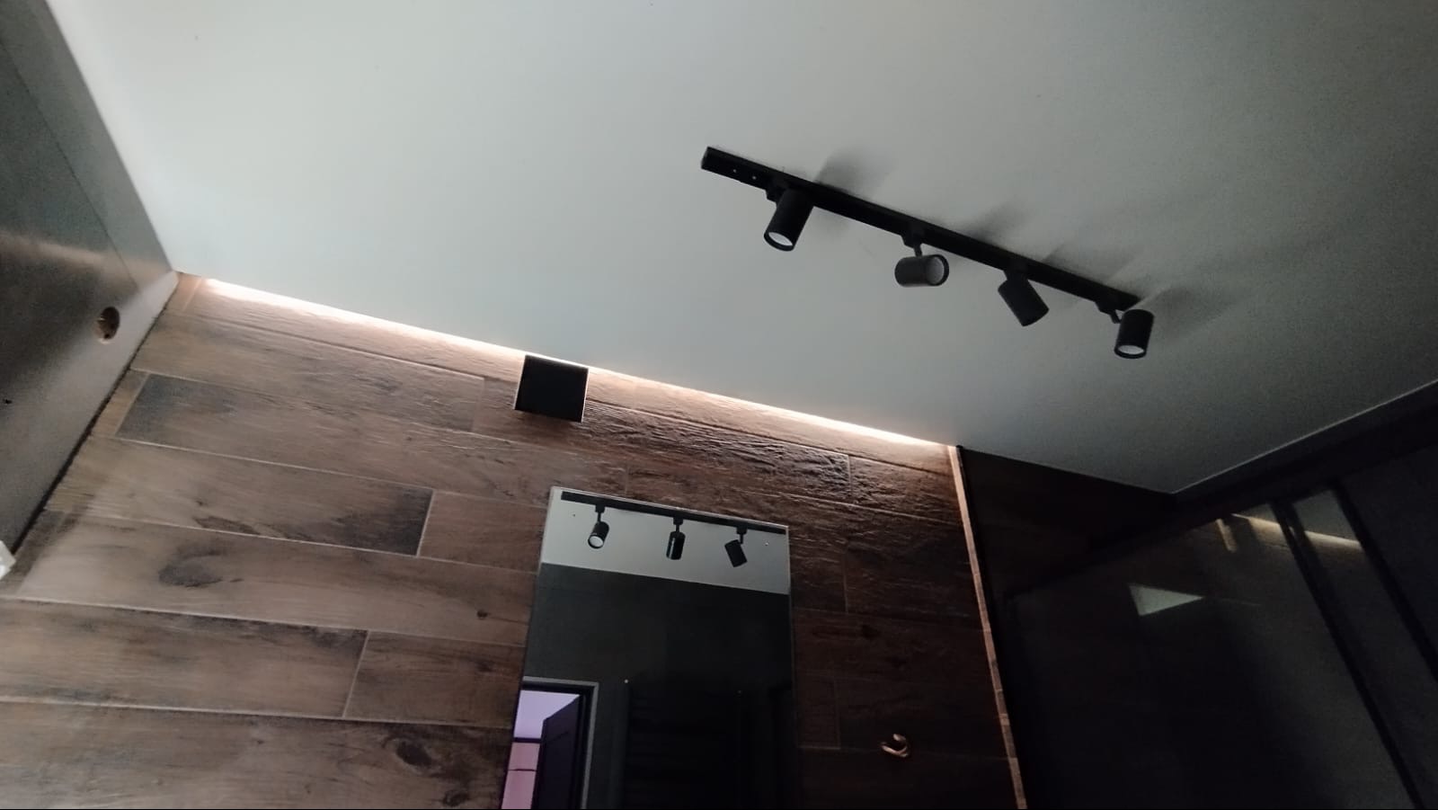 Nowoczesna łazienka z drewnopodobnymi płytkami, oświetleniem punktowym na suficie i podświetleniem LED wzdłuż ściany. Lustro z dodatkowym oświetleniem.