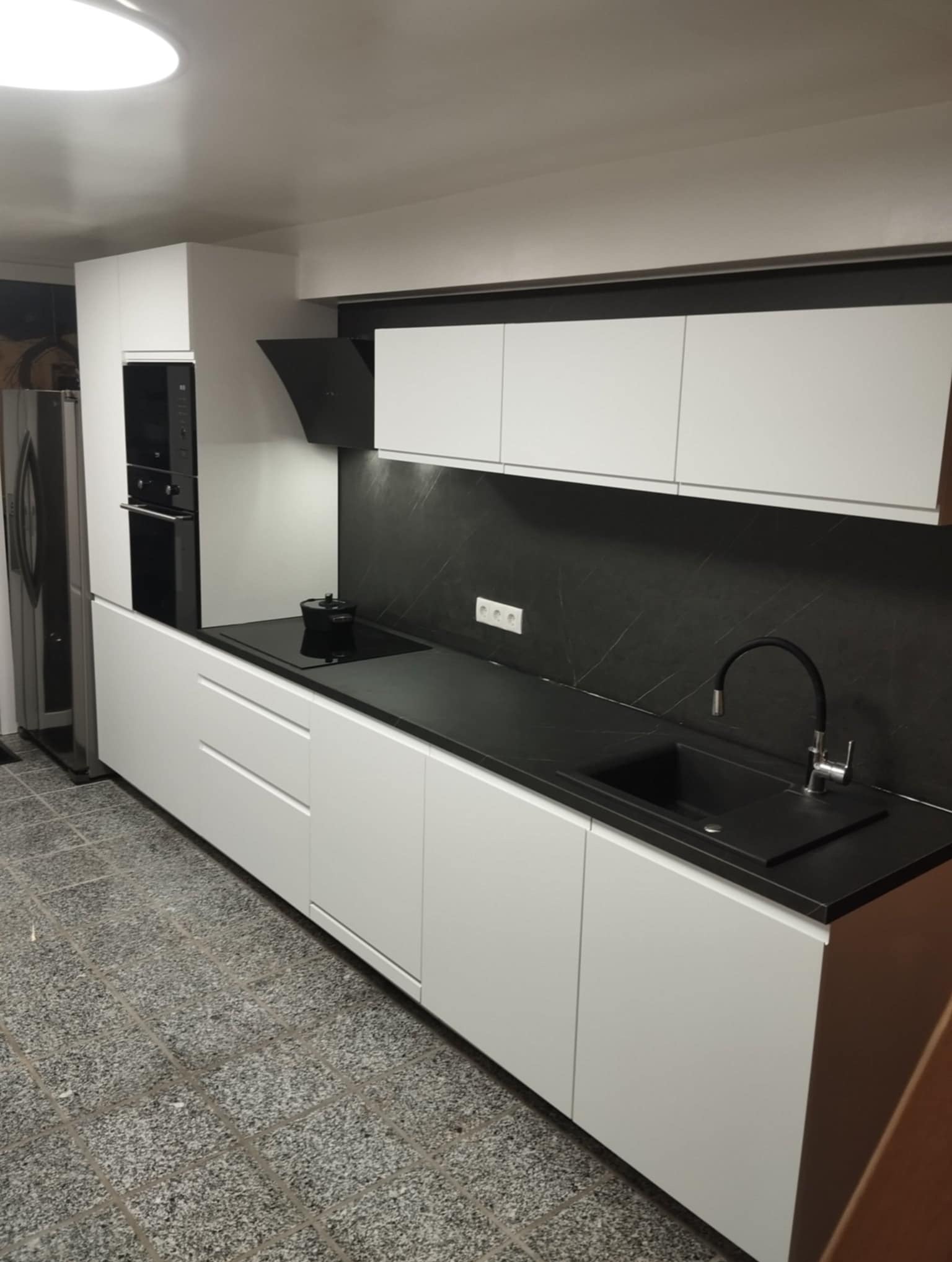 Nowoczesna kuchnia z białymi szafkami i czarnym blatem, minimalistyczny design, czarny panel nad blatem, widok na zlew i płytę grzewczą.