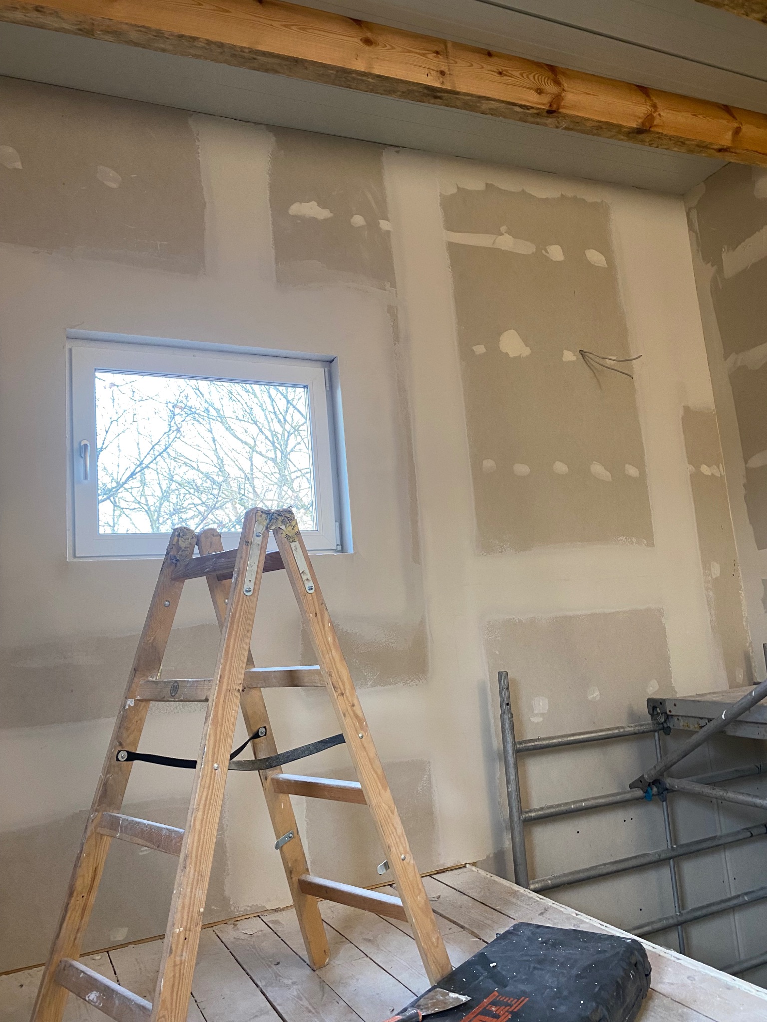Ściana z płyt gipsowo-kartonowych w trakcie szpachlowania, widoczne łączenia i drabina. Surowe drewniane belki stropowe dodają rustykalnego charakteru.