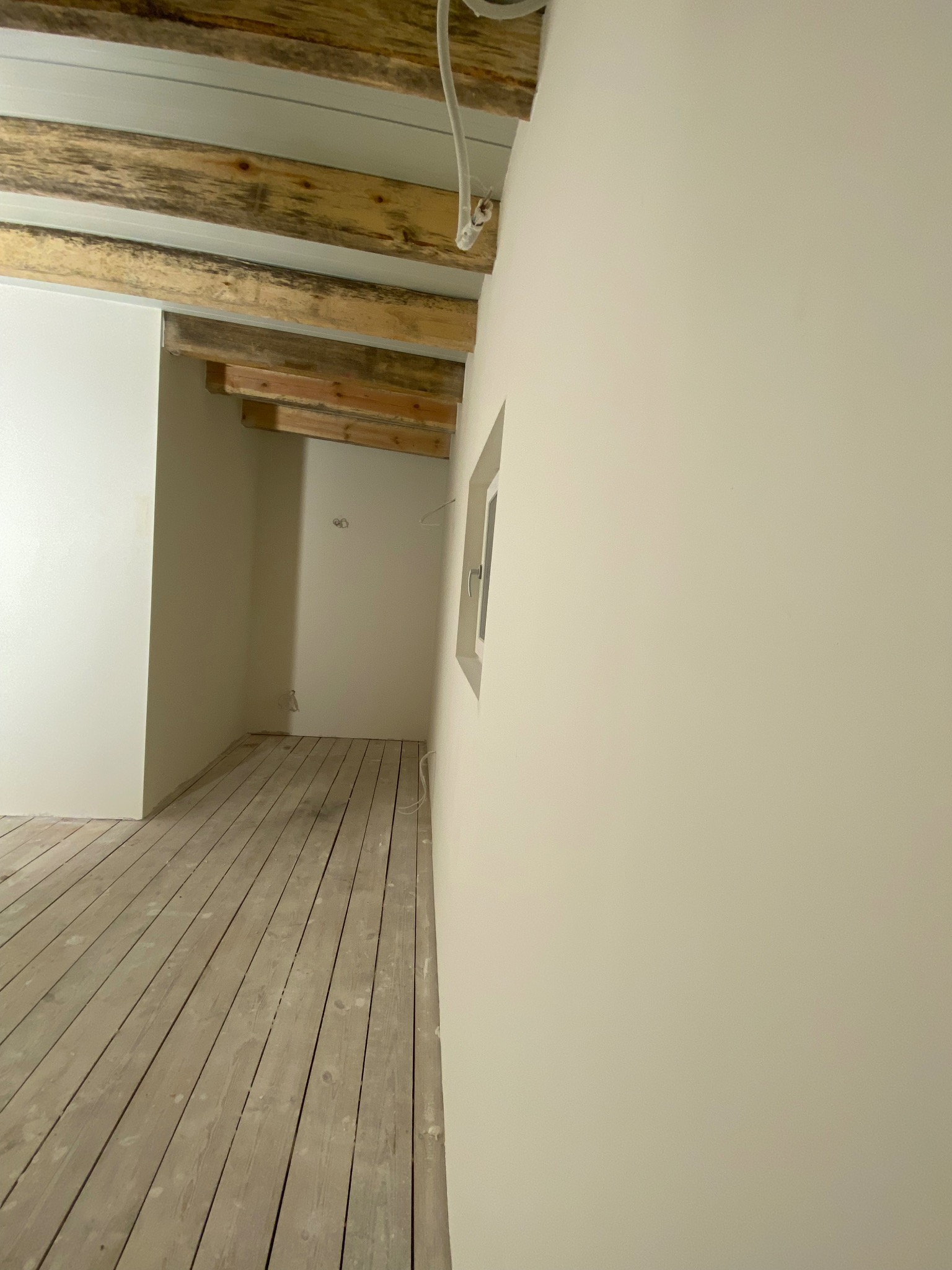 Jasne wnętrze po odświeżeniu: pomalowane ściany, drewniana podłoga i belki stropowe. Widoczne okno i instalacja elektryczna.