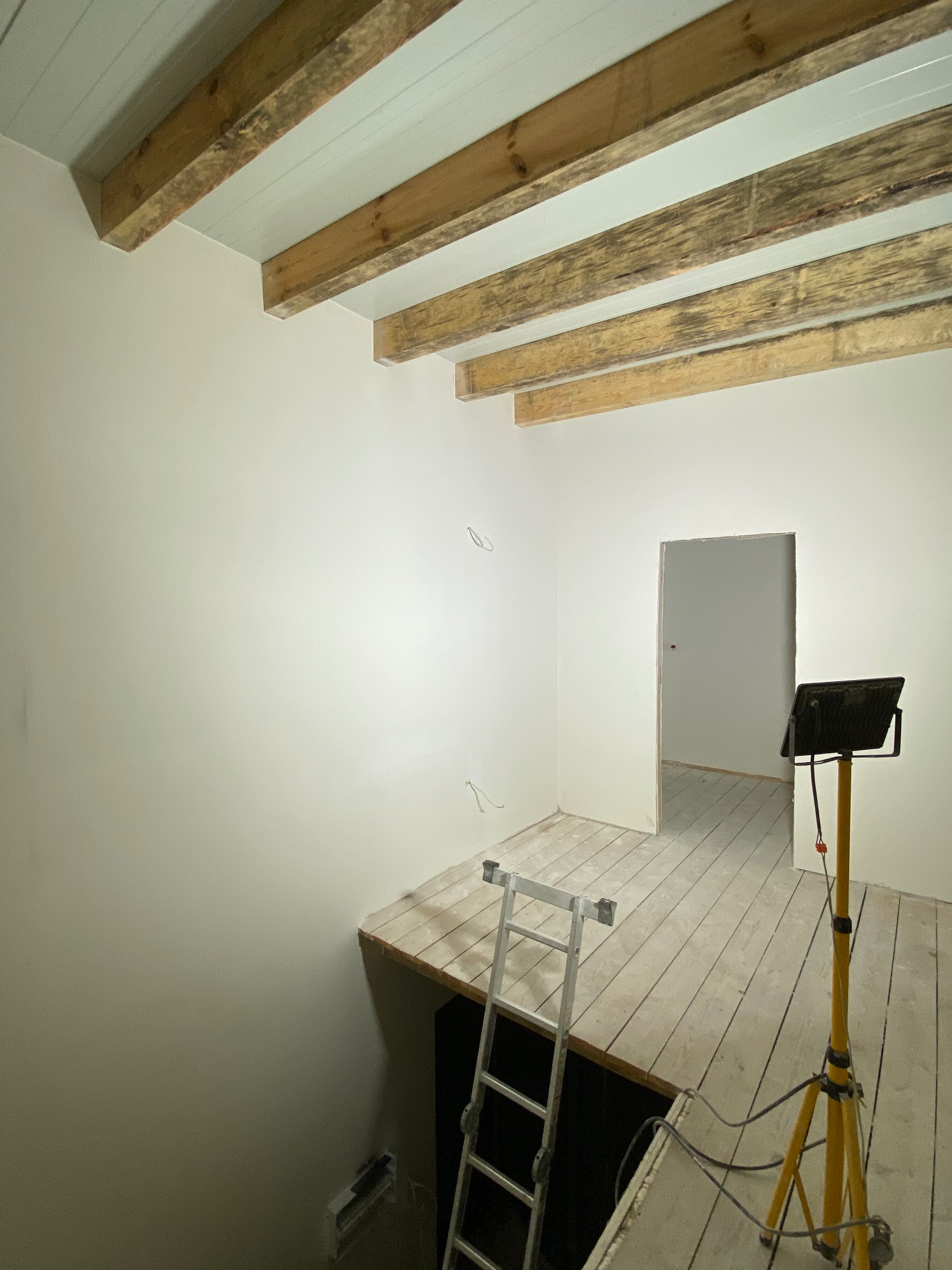 Wnętrze w trakcie prac: białe ściany, drewniane belki stropowe, surowe deski podłogowe, drabina i lampa budowlana. Widoczne kable i otwór w podłodze.