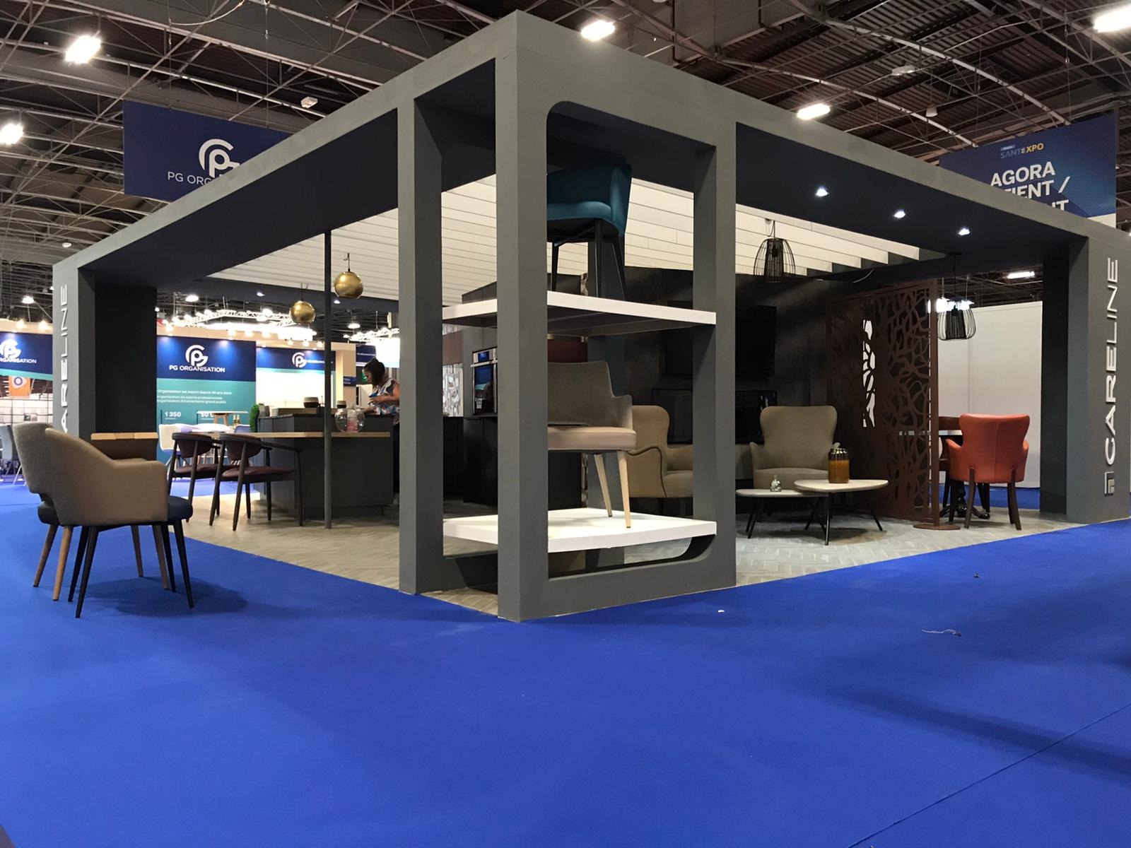 Stoisko targowe z szarą ramą, prezentujące aranżacje meblowe na różnych poziomach, z krzesłami w odcieniach beżu, turkusu i pomarańczy oraz niebieską wykładziną podłogową.