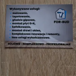 Wizytówka firmy budowlanej FOR-BUD leżąca na drewnianej powierzchni. Wizytówka wymienia usługi takie jak malowanie, tapetowanie, gładzie gipsowe, montaż płyt G-K, kafelkowanie, montaż drzwi i okien...