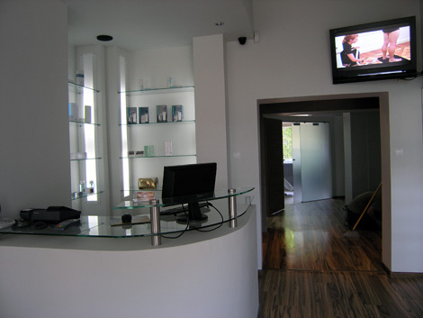 Recepcja w salonie kosmetycznym z szklanym blatem, komputerem, kasą fiskalną, regałami z produktami, telewizorem pokazującym zabieg depilacji laserowej i widokiem na korytarz z przeszklonymi drzwiami.