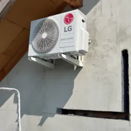 Biała jednostka zewnętrzna klimatyzacji LG Dual Inverter zamontowana na wspornikach na szarej ścianie budynku, widoczny fragment drabiny i drewniana konstrukcja dachu