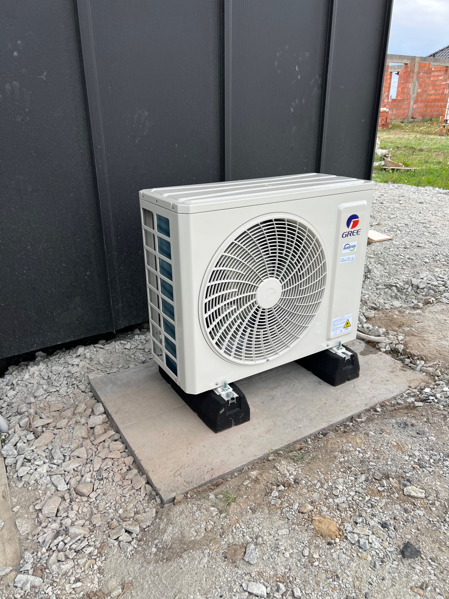 Zewnętrzna jednostka klimatyzacji marki Gree, zamontowana na betonowej podstawie z gumowymi podkładkami tłumiącymi drgania, na tle czarnej ściany budynku i placu budowy.