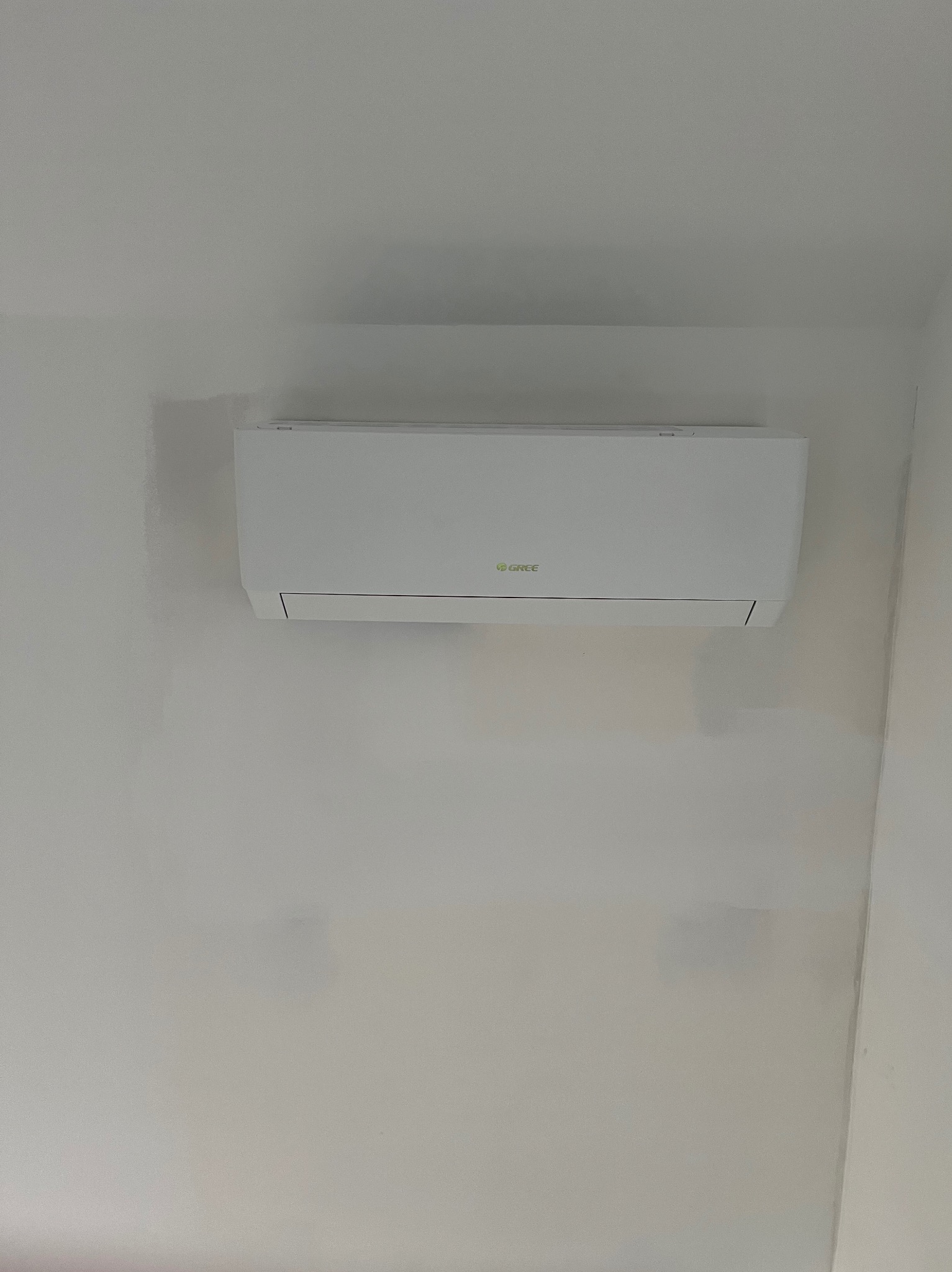 Biały klimatyzator marki Gree zamontowany na jasnej ścianie, widoczne ślady po malowaniu.