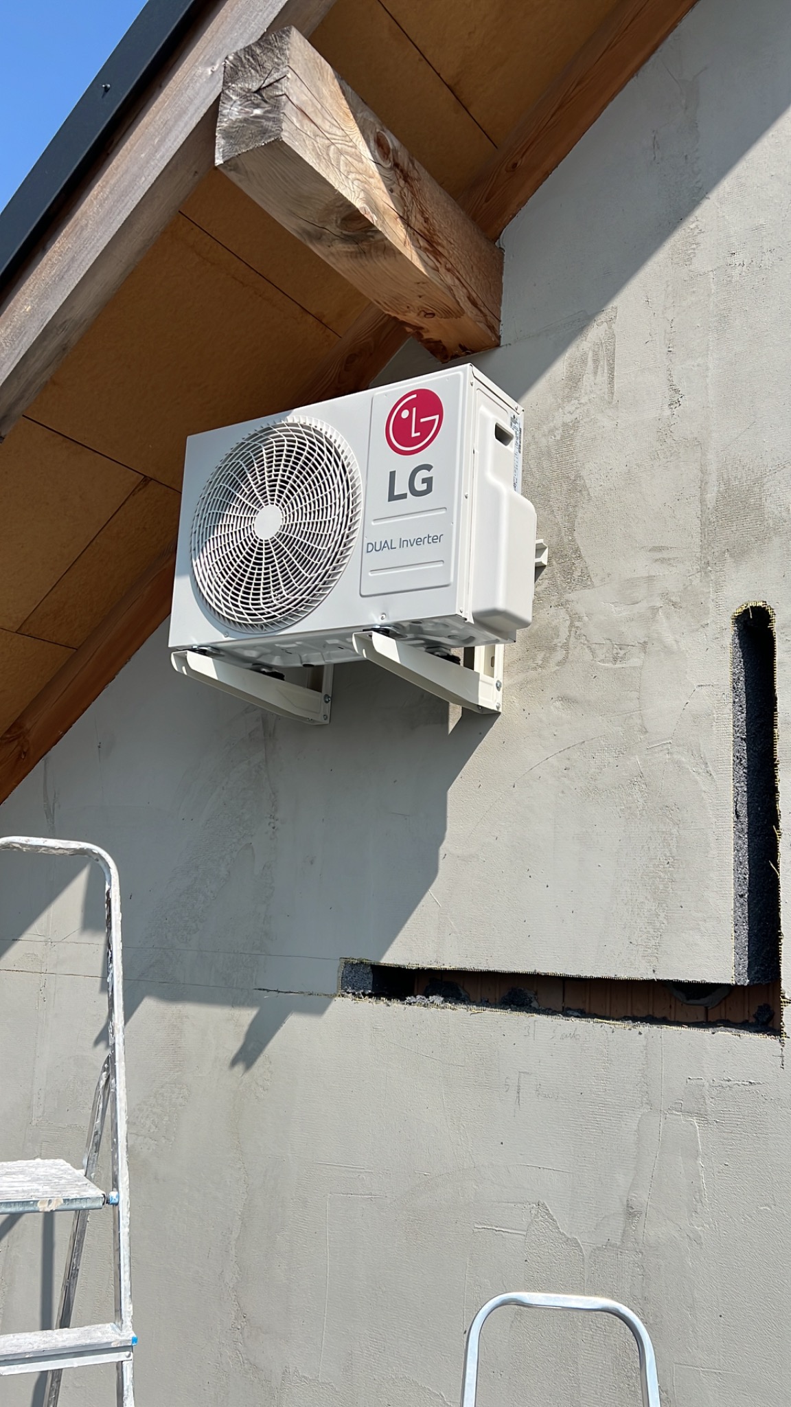 Biała jednostka zewnętrzna klimatyzacji LG Dual Inverter zamontowana na wspornikach na szarej ścianie budynku, widoczny fragment drabiny i drewniana konstrukcja dachu