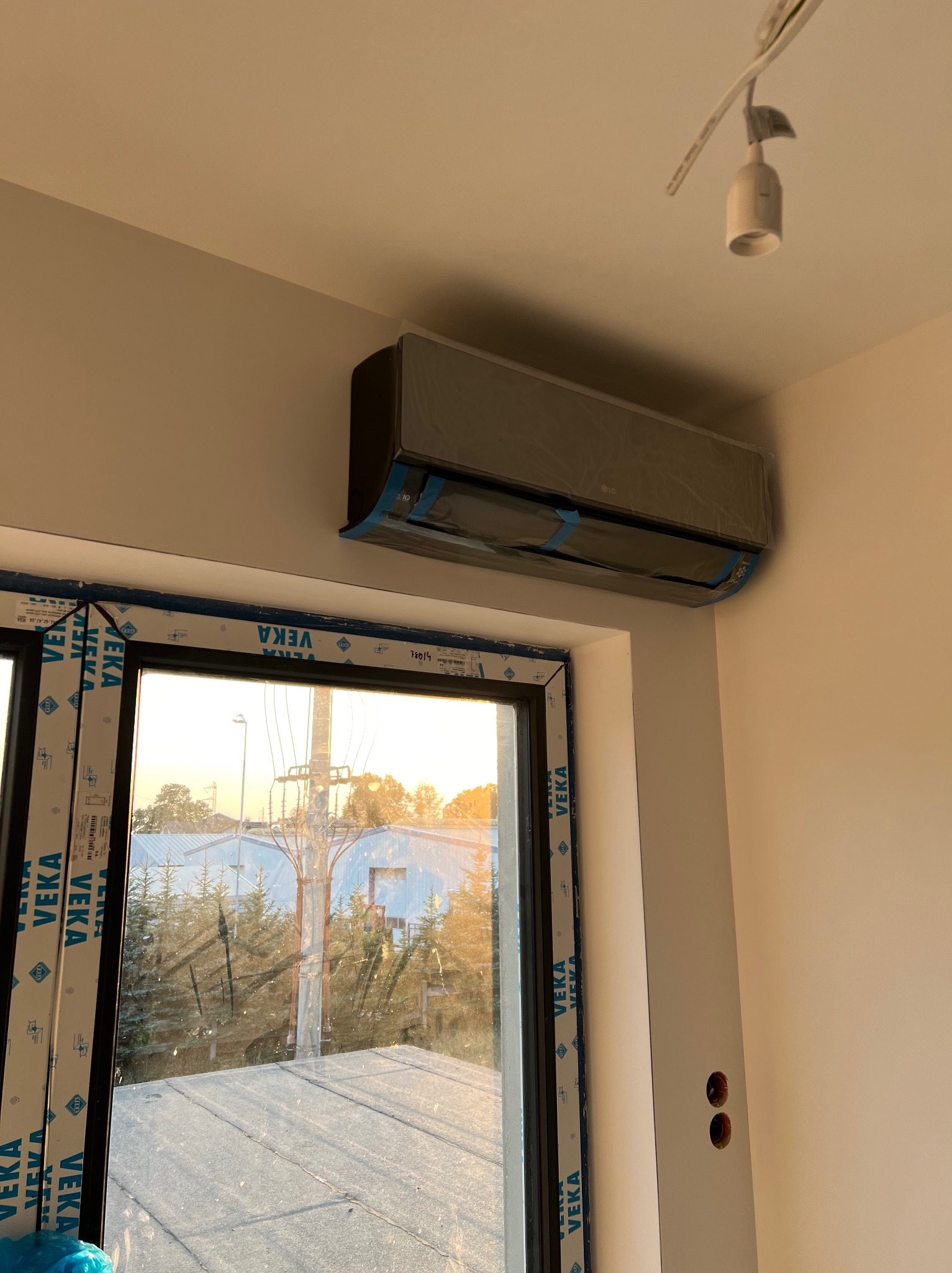 Nowo zamontowana jednostka klimatyzacyjna LG, jeszcze w folii ochronnej, w jasnym pomieszczeniu z widokiem na zewnątrz przez okno z ramą VEKA.