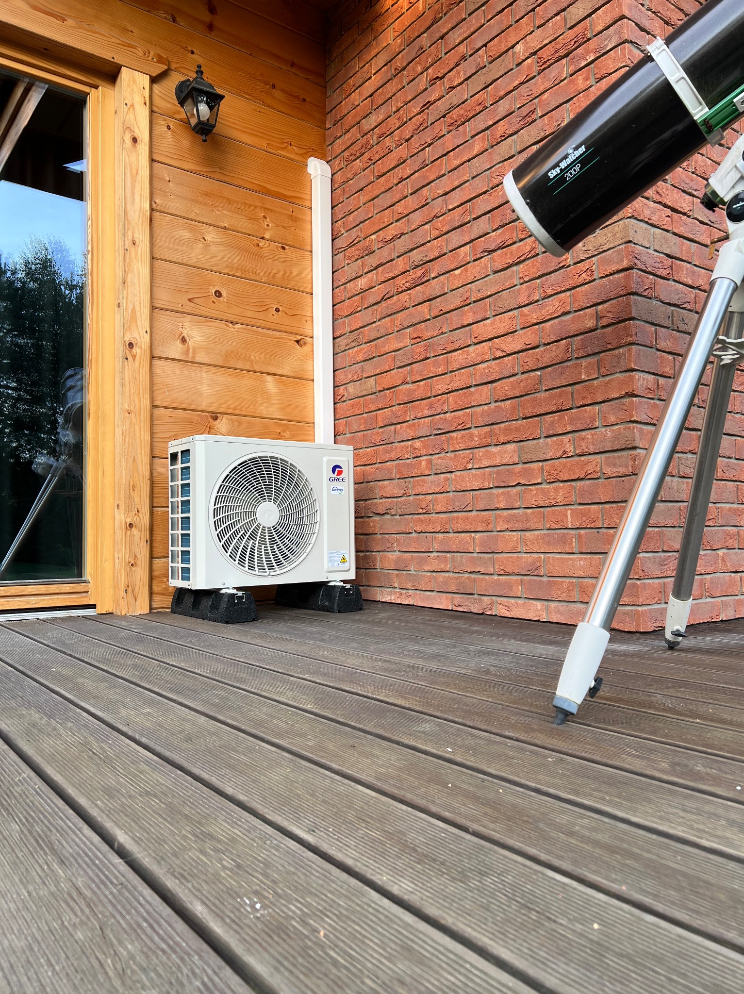 Zewnętrzna jednostka klimatyzacji marki Gree umieszczona na czarnych podkładkach na drewnianym tarasie, obok ściany z czerwonej cegły i teleskopu Sky-Watcher 200P.