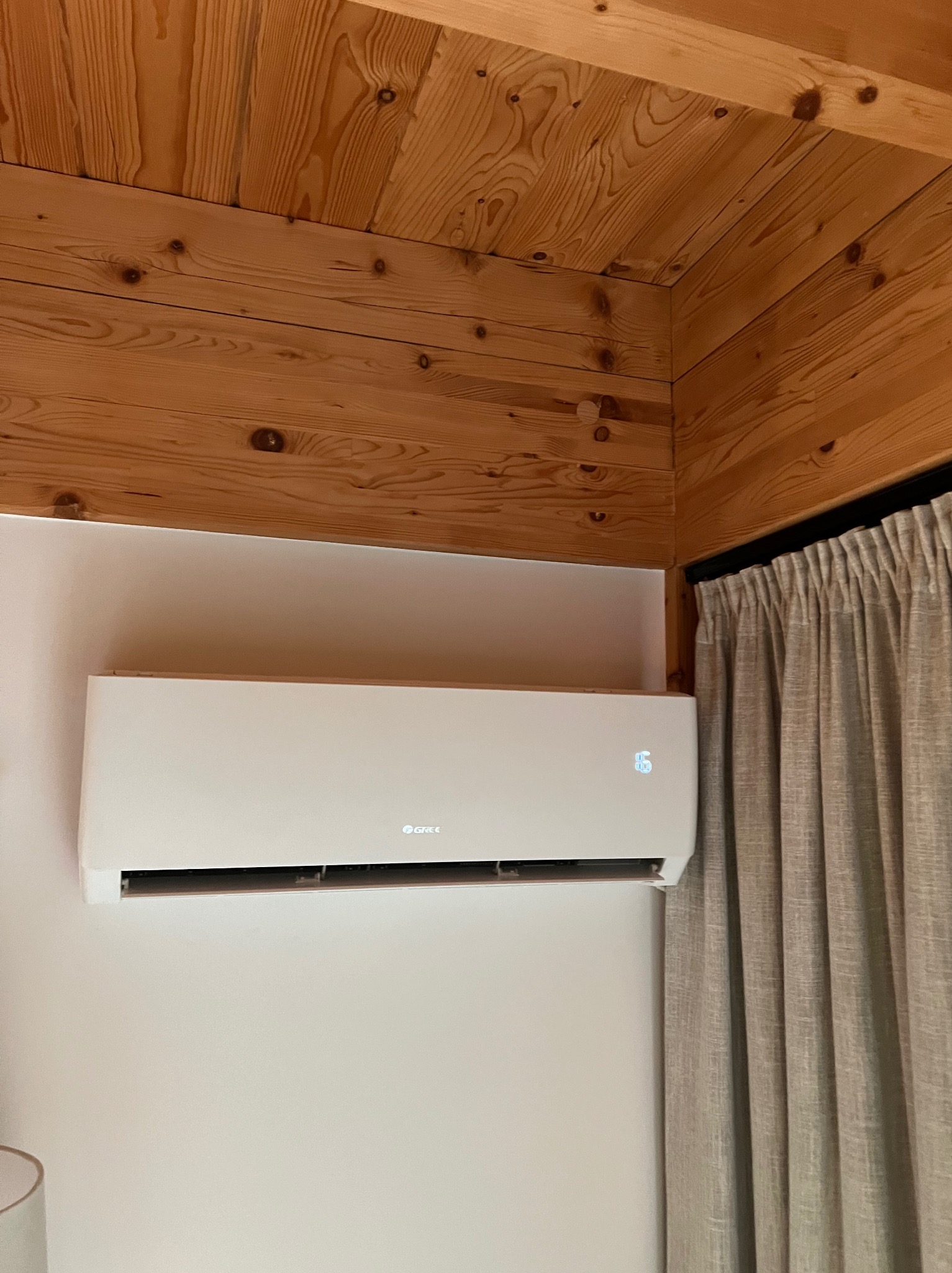 Biały klimatyzator marki Gree zamontowany na ścianie w pomieszczeniu z drewnianym sufitem i zasłonami w tle.