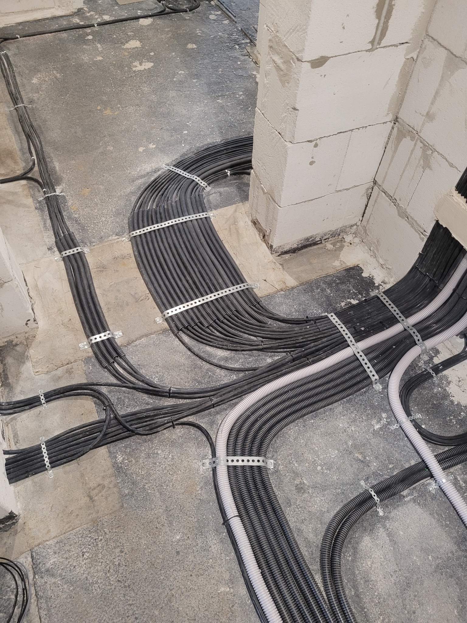 Wiązki czarnych i szarych peszli elektrycznych, mocowanych metalowymi uchwytami do surowego betonowego podłoża w nowo budowanym budynku z białych bloczków.