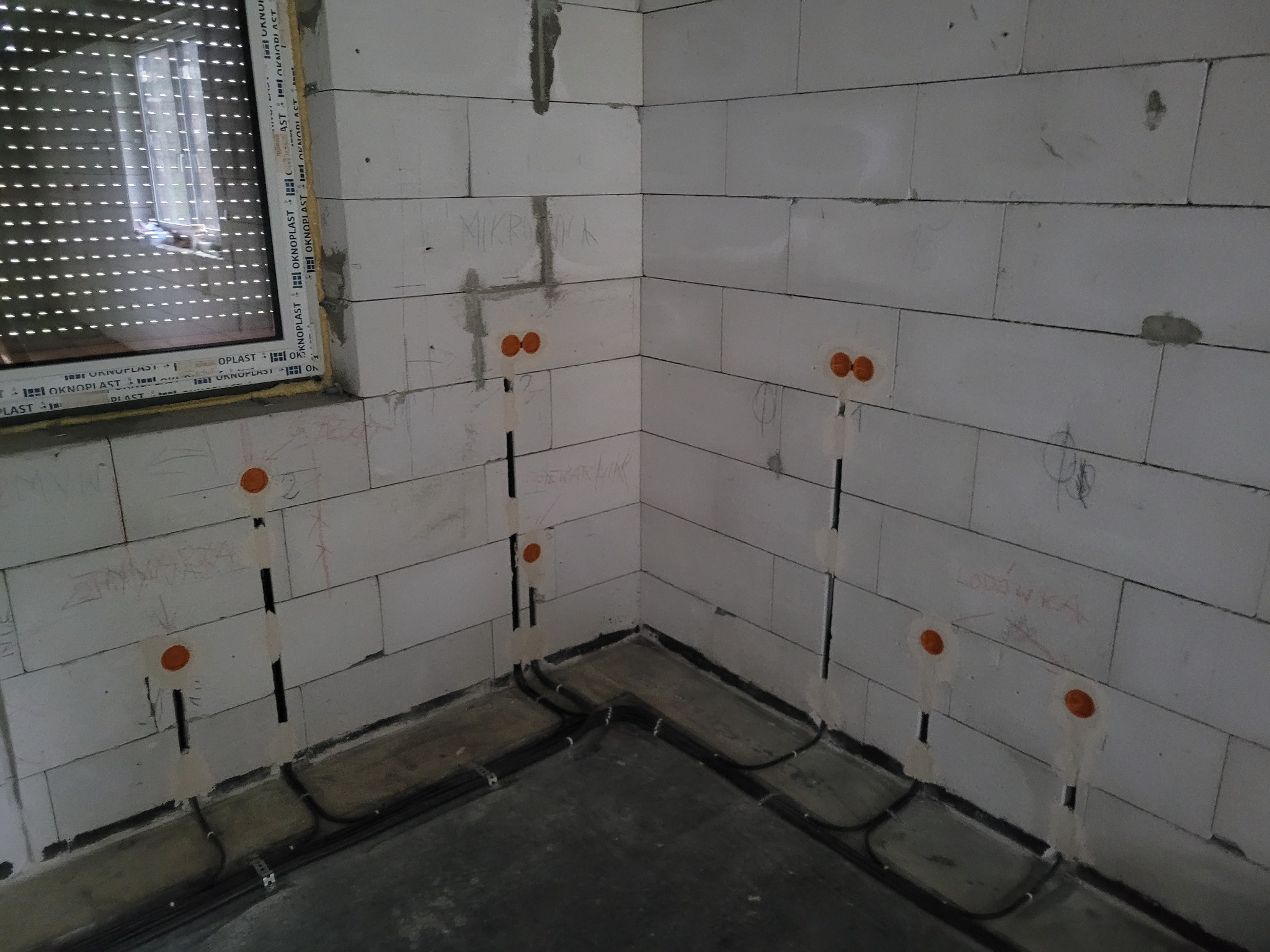 Wewnętrzna instalacja elektryczna w surowym pomieszczeniu z betonu komórkowego, widoczne przewody w bruzdach i puszki instalacyjne, okno z żaluzjami w tle.