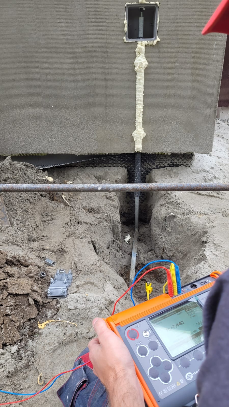 Pomiar uziemienia instalacji odgromowej w wykopie, widoczny miernik elektryczny Sonel z podłączonymi przewodami pomiarowymi, metalowa bednarka i rura z pianką montażową przy ścianie budynku.