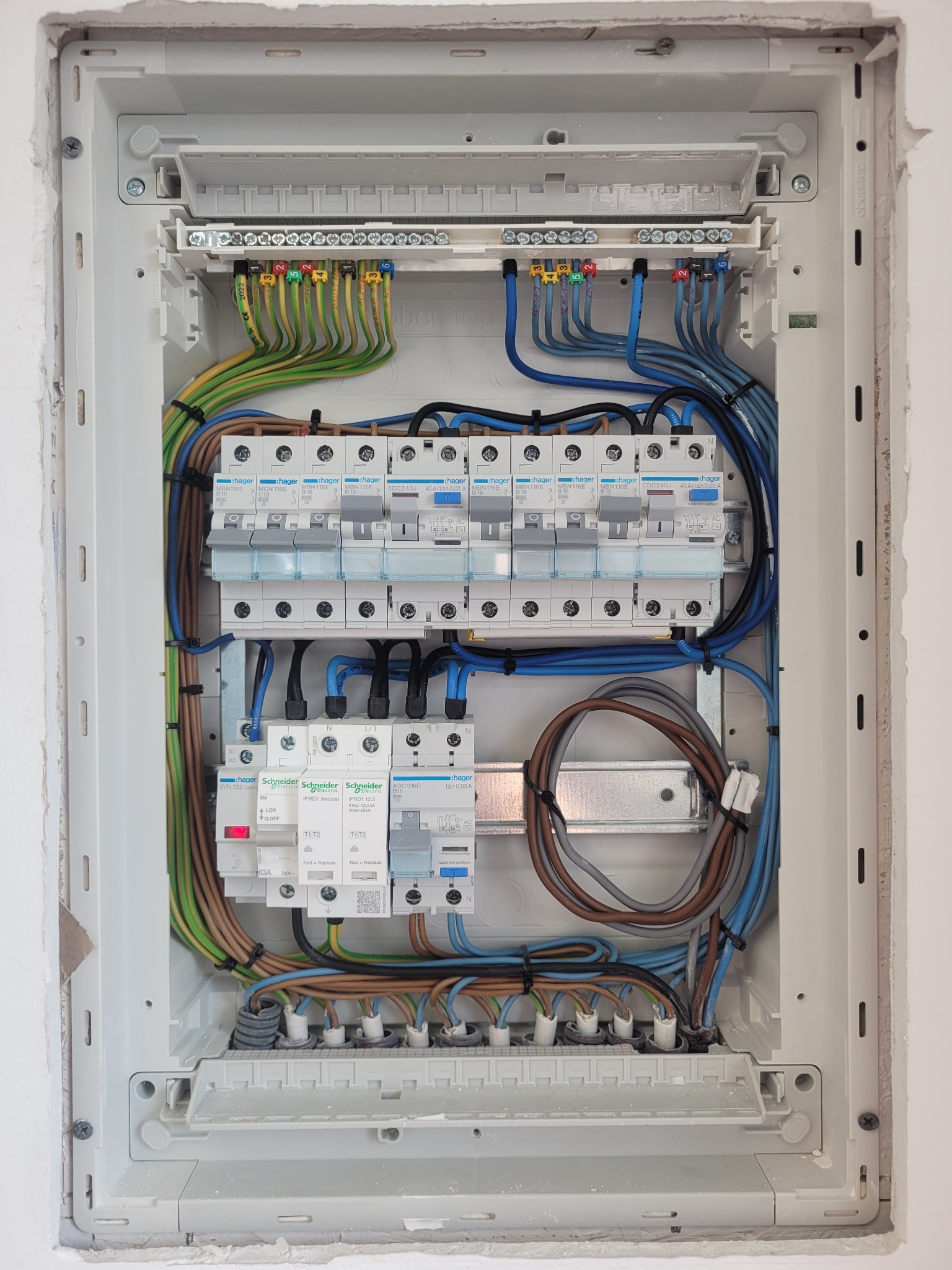 Otwarta skrzynka bezpiecznikowa Hager z widocznymi przewodami oznaczonymi kolorami i numerami, zamontowana w ścianie z surowym wykończeniem.
