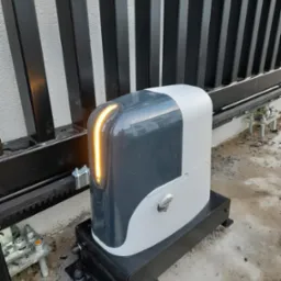 Szary automat do bramy przesuwnej z podświetleniem LED, zamontowany na czarnej podstawie przy metalowej bramie z pionowymi szczeblami i betonowym podłożem.
