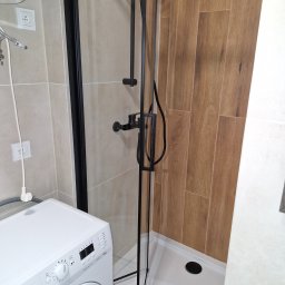 Pawex-bud - Nowoczesna łazienka z kabiną prysznicową typu walk-in z czarnymi elementami, drewnopodobne płytki na ścianie, biała pralka z przodu, bojler nad pralką.