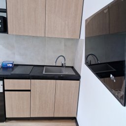 Pawex-bud - Nowoczesna, jasna kuchnia z drewnopodobnymi frontami szafek, szarym blatem i zlewozmywakiem oraz płytą grzewczą. W odbiciu na ścianie widoczny fragment tego samego blatu i zlewu.