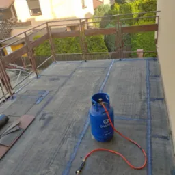 Niedokończona izolacja dachu płaskiego z widoczną butlą gazową, palnikiem i fragmentami papy na tle zieleni ogrodowej i zabudowy jednorodzinnej.