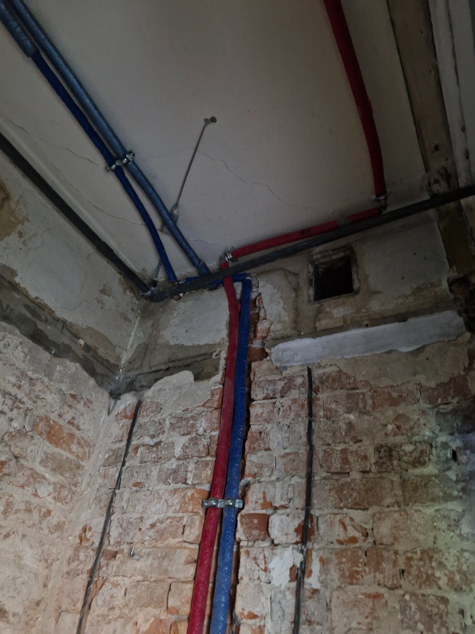 Narożnik pomieszczenia w trakcie remontu z odsłoniętą cegłą, widoczne rury w izolacji czerwonej i niebieskiej, przymocowane metalowymi uchwytami do ściany i sufitu.