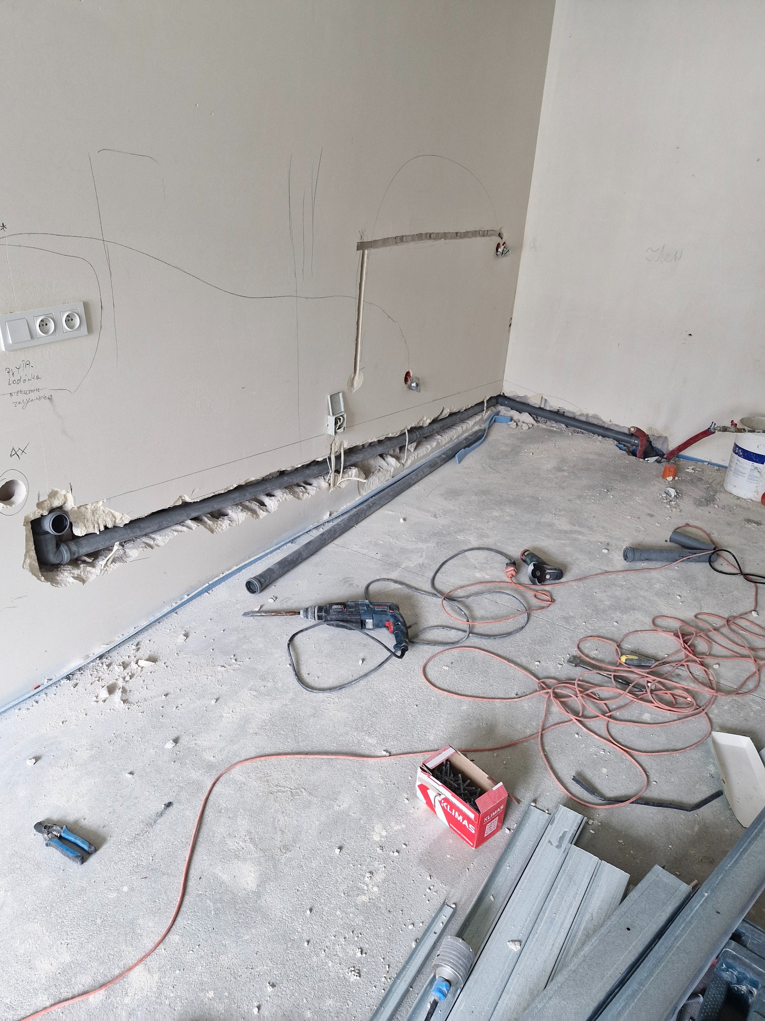 Wnętrze w trakcie prac instalacyjnych: widoczne przewody elektryczne, rury kanalizacyjne w bruzdach ściennych, narzędzia budowlane i profile metalowe na podłodze.