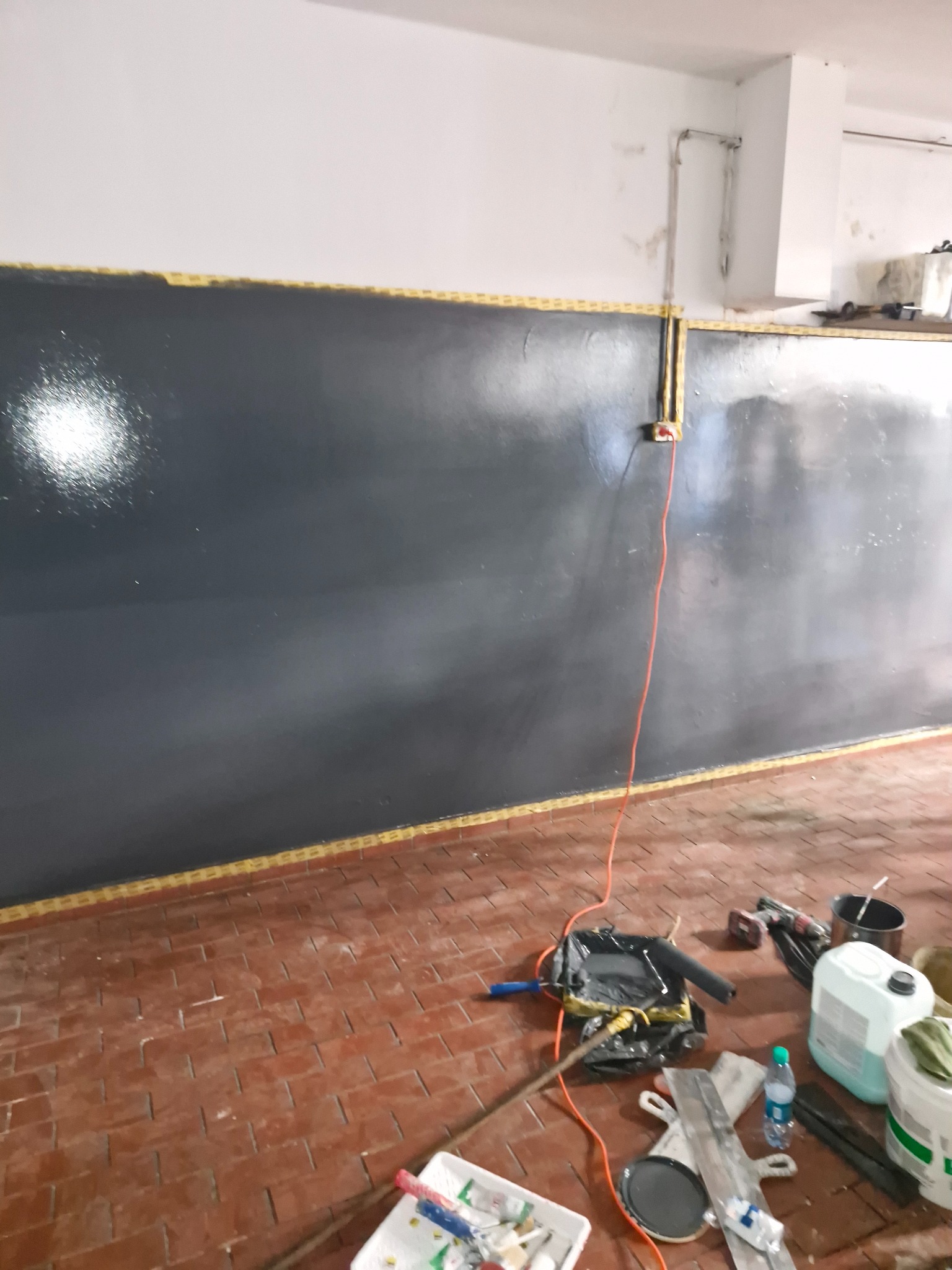 Świeżo pomalowana ciemnoszara ściana z żółtą taśmą malarską na górze, widoczne narzędzia malarskie i akcesoria na ceglanej podłodze, w tym wiadro z farbą, wałki i butelka wody.
