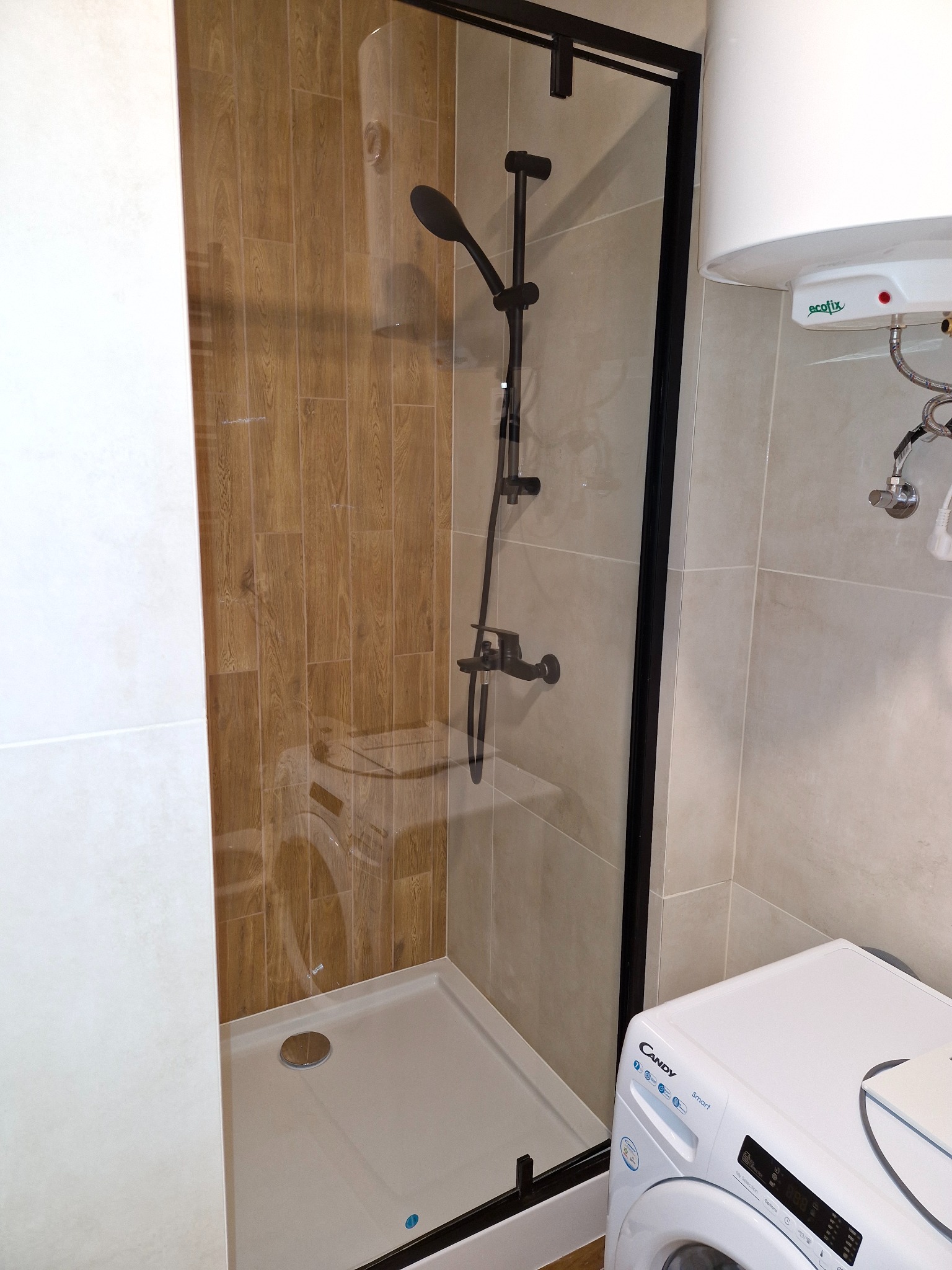 Nowoczesna kabina prysznicowa z czarną armaturą na tle płytek imitujących drewno, obok pralka Candy i bojler Ecofix.