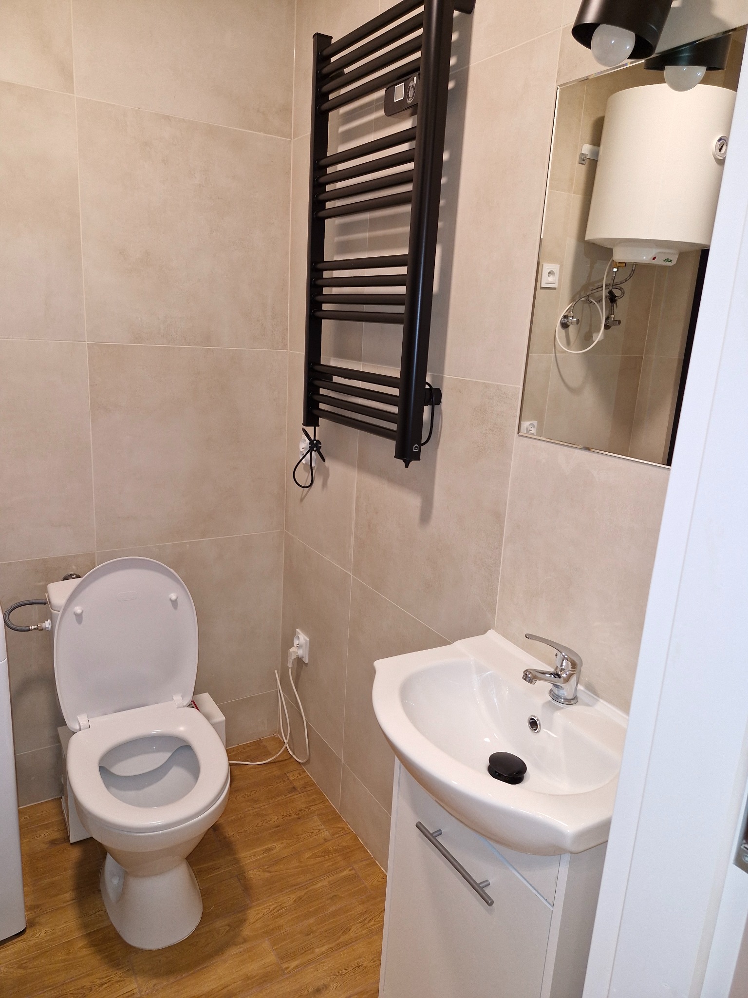 Nowoczesna, jasna łazienka z beżowymi płytkami, drewnopodobną podłogą, białym zlewem z szafką, wiszącym grzejnikiem drabinkowym i toaletą z otwartą klapą.