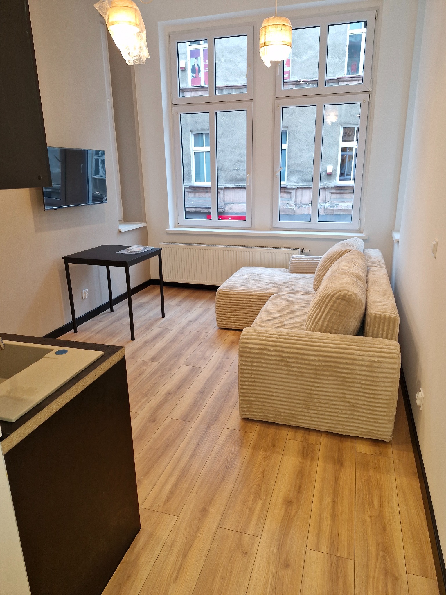 Jasny pokój z drewnianą podłogą, beżową sofą z poduszkami, czarnym stolikiem i widokiem z okna na kamienicę, lampy wiszące z abażurami.