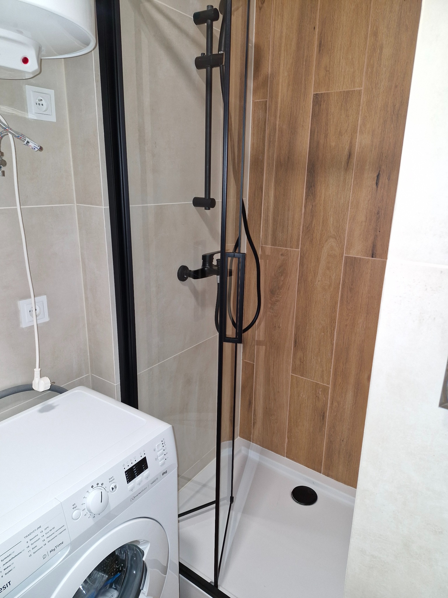Nowoczesna łazienka z kabiną prysznicową typu walk-in z czarnymi elementami, drewnopodobne płytki na ścianie, biała pralka z przodu, bojler nad pralką.